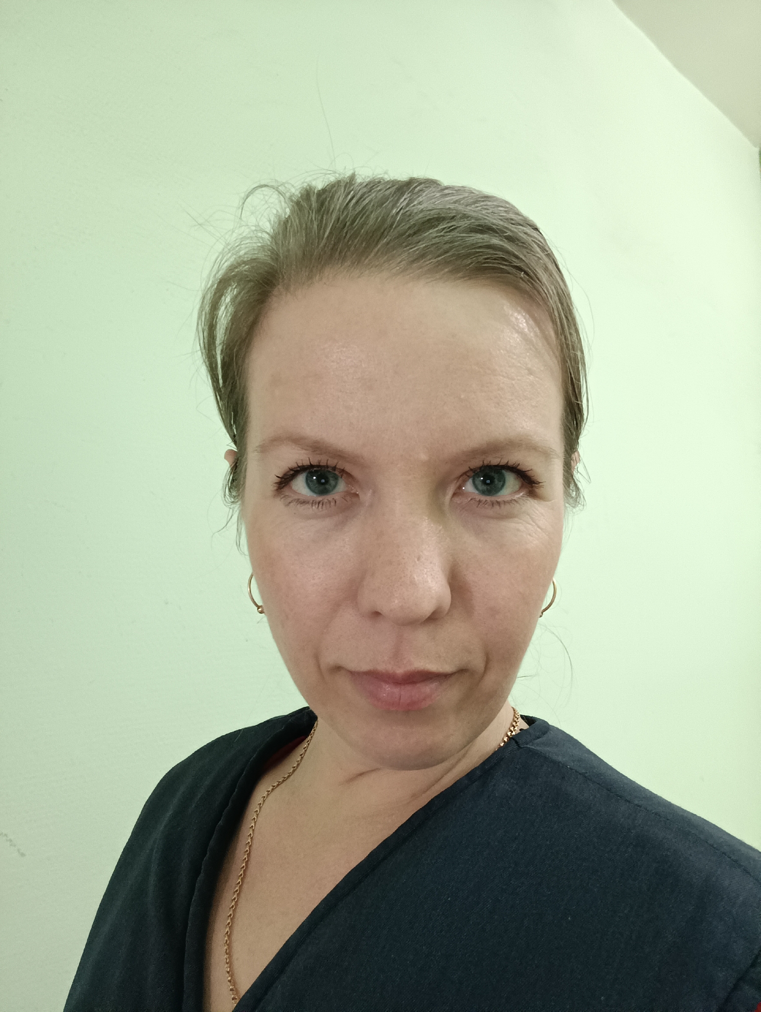 A close-up headshot of a fair-skinned woman with graying blonde hair pulled back, wearing a dark-colored top and gold earrings and necklace. She is looking directly at the camera with blue eyes. The background is a light green wall. The photograph may have been taken indoors. There is no information to indicate the location is in Chaikovsky, Russia.
