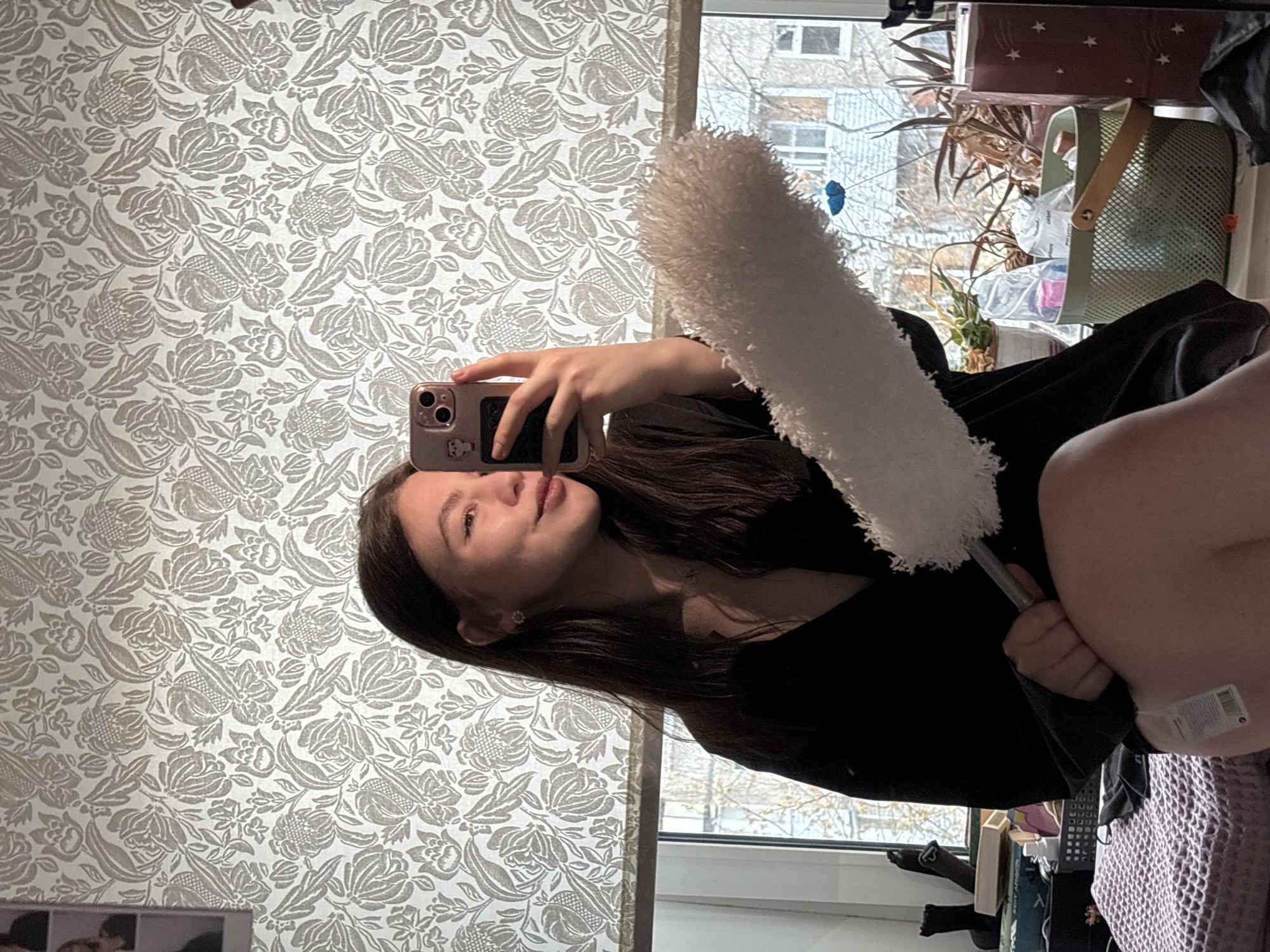 A young woman is captured in a mirror selfie, holding a white fluffy duster prominently in the upper right of the frame (when correctly oriented). She has long dark hair and is wearing a dark top, with a calm, slightly smiling expression as she holds a smartphone, clad in a light-colored case with small stickers, to take the picture.
The background to her left (in the reflection) showcases a wall adorned with light grey patterned wallpaper featuring a floral or foliage design. To her right, a window offers a view of the outdoors, where bare trees and a multi-story residential building are visible under a bright, diffused sky, typical of daylight on an overcast day. This architectural style and the seasonal appearance of the trees are consistent with urban environments in Perm, Russia.
The window sill below is arranged with several items, including potted plants (one with broad leaves, another smaller), a green mesh basket containing plastic bags and other miscellaneous items, and a reddish-brown box decorated with stars. In the lower right corner, a textured fabric, possibly a purple or lavender blanket, is partially visible, alongside a white product tag displaying a barcode and small, illegible text. The scene conveys a casual, domestic moment, possibly related to tidying up.