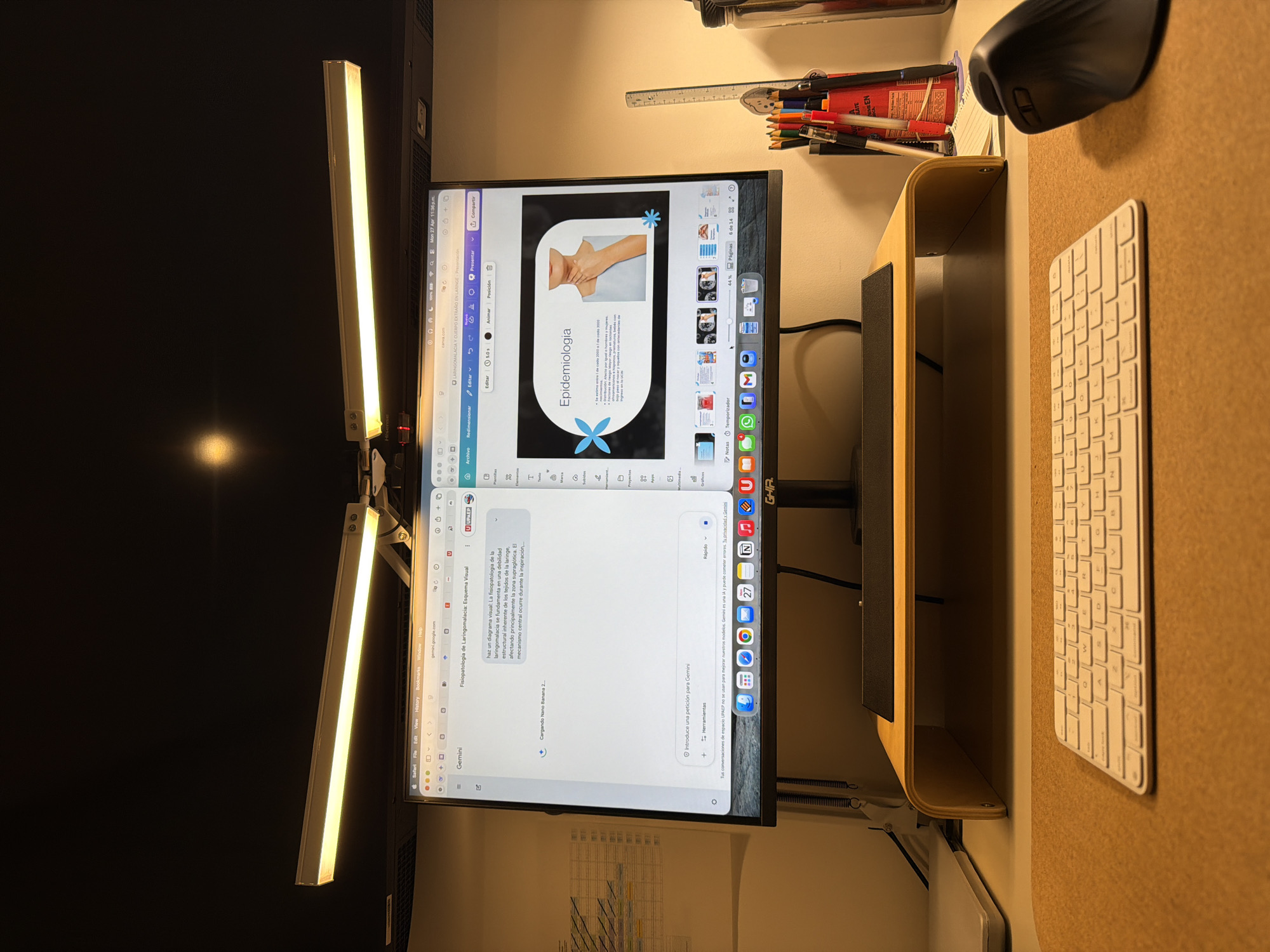 This media file captures a late-night desk setup in Santa Clara Ocoyucan, Mexico, suggesting focused work or study. The central subject is a large computer monitor, positioned vertically (portrait mode), displaying two active applications.
The top application is Canva, showing a presentation slide titled "Epidemiología" (Epidemiology). The slide features an image of a person's neck and chest, along with Spanish text discussing epidemiology and two light blue snowflake/starburst graphics. The top bar of this application indicates "Mon 27 Apr 11:36 p.m." and "U UPAEP," likely referencing a university.
Below, the monitor displays Google Gemini, an AI assistant. This window contains Spanish text about "Fisiopatología de Laringomalacia: Esquema Visual" (Pathophysiology of Laryngomalacia: Visual Scheme), detailing the structural weakness in the larynx. A prompt reads, "Introduce una petición para Gemini" (Introduce a request for Gemini). The monitor's dock, visible at the bottom, shows various application icons including Safari, Mail, Calendar (with '27'), and Notes.
Two long, warm-toned LED light bars are attached to the top of the monitor, illuminating the workspace. To the right of the monitor, on a light brown, textured desk mat, sits a white Apple-style keyboard and a black ergonomic Logitech mouse. The monitor rests on a simple, light wood-colored riser. In the upper right background, a pencil holder filled with pens and pencils, along with a ruler, is visible. The overall scene suggests a dedicated study or work session, likely for academic or professional purposes given the content on screen, occurring late in the evening.
