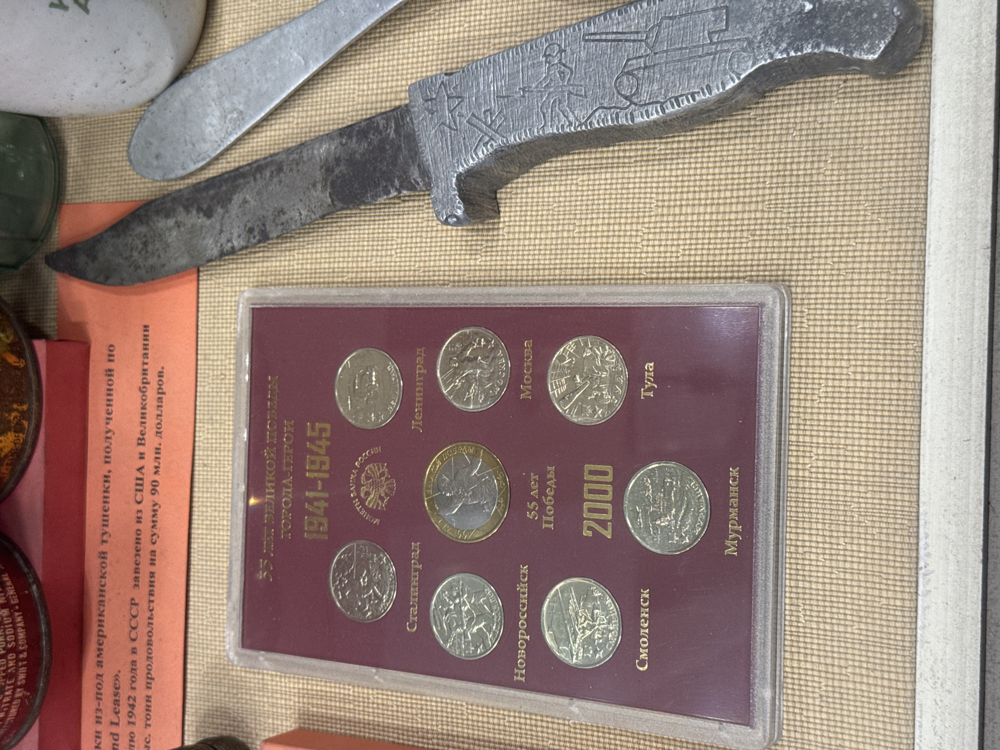 The image displays a collection of objects, including a knife with etched military imagery, a set of commemorative coins in a maroon-colored case, and a page of text in Russian. The knife, positioned diagonally across the top left, appears to be made of metal and features engravings of military figures and vehicles on its handle. Below the knife, the coin set commemorates the "City Heroes" of the Great Victory (World War II), with coins labeled with city names such as Leningrad, Moscow, Tula, Murmansk, and others. A document with Russian text is partially visible, referencing "American stew" and aid received from the USA and Great Britain. The scene is in Saint Petersburg, Russia.