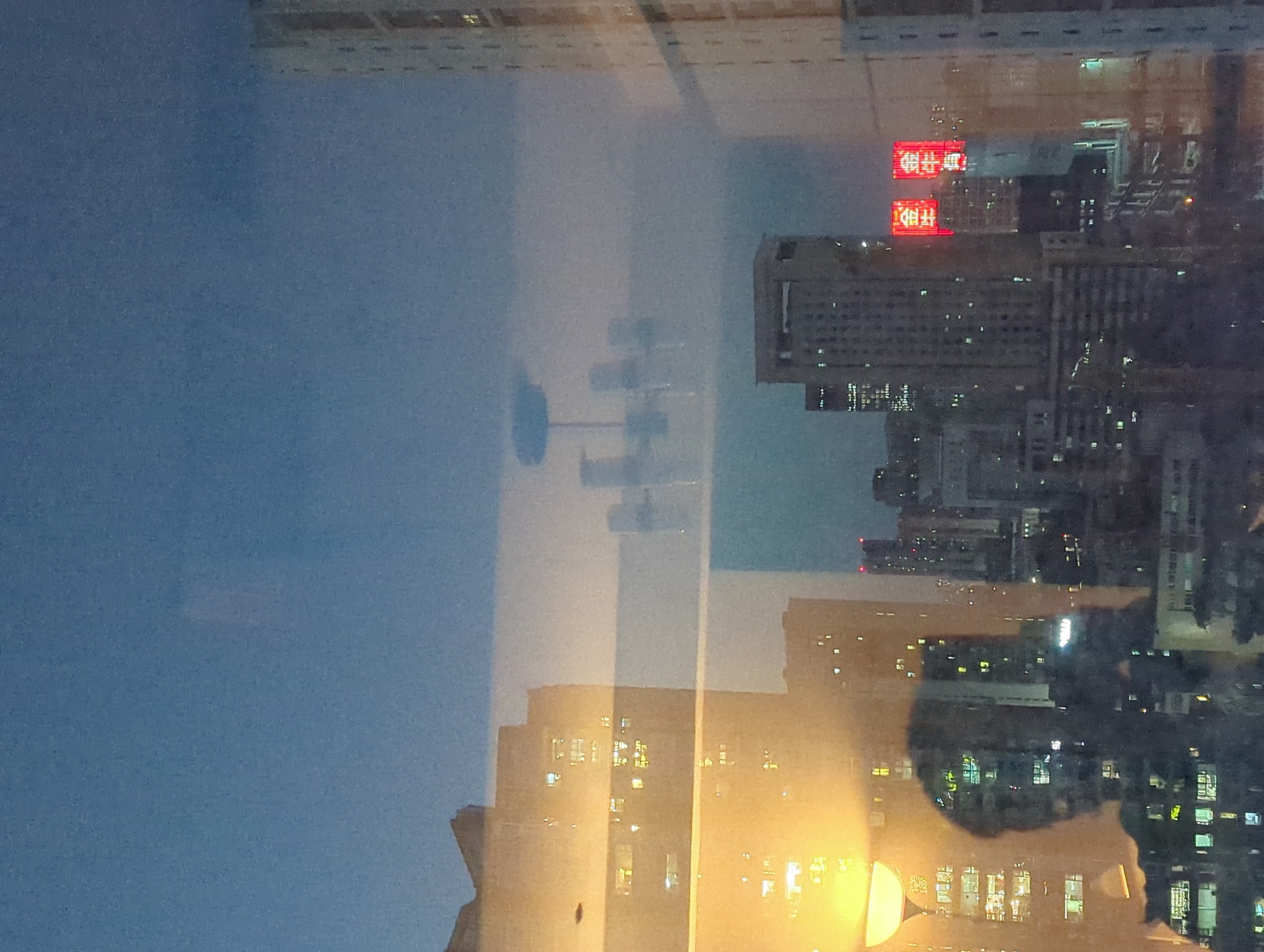The image captures a nighttime city view through a window, likely from a high-rise building in Qingyang District, China. The scene is dominated by a reflection on the glass, which partially obscures the view of the city skyline.
In the foreground, a reflection of a person, likely the photographer, is visible as a dark silhouette against the illuminated interior of a room, suggested by a large, warm-toned lamp on the left. The reflection shows the person holding a camera.
Beyond the reflection, the city skyline features numerous tall buildings with illuminated windows, indicating a densely populated urban area. In the middle ground, a chandelier-like object is reflected, appearing as a hazy, multi-armed structure. Further into the distance, two prominent red neon signs with Chinese characters are visible, glowing brightly against the darker buildings. These signs add a distinct visual element to the urban landscape. The sky above is a deep blue, transitioning to a lighter hue towards the horizon, suggesting dusk or early night. The overall impression is one of a bustling, illuminated city at night, viewed from an indoor vantage point.