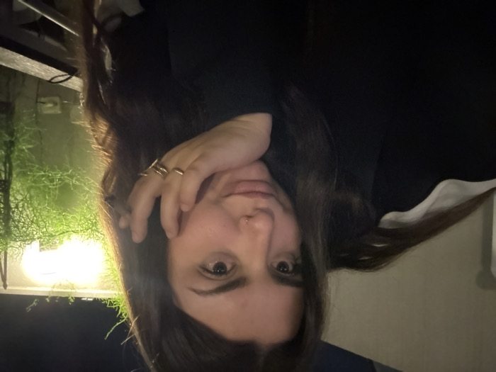 A woman with long brown hair and fair skin is facing the camera. Her hand is near her face, displaying several rings on her fingers. She wears a dark top with a glimpse of a white collar. The background shows a dimly lit outdoor space, including greenery illuminated by a light, suggesting a nighttime setting. The photo is slightly tilted.