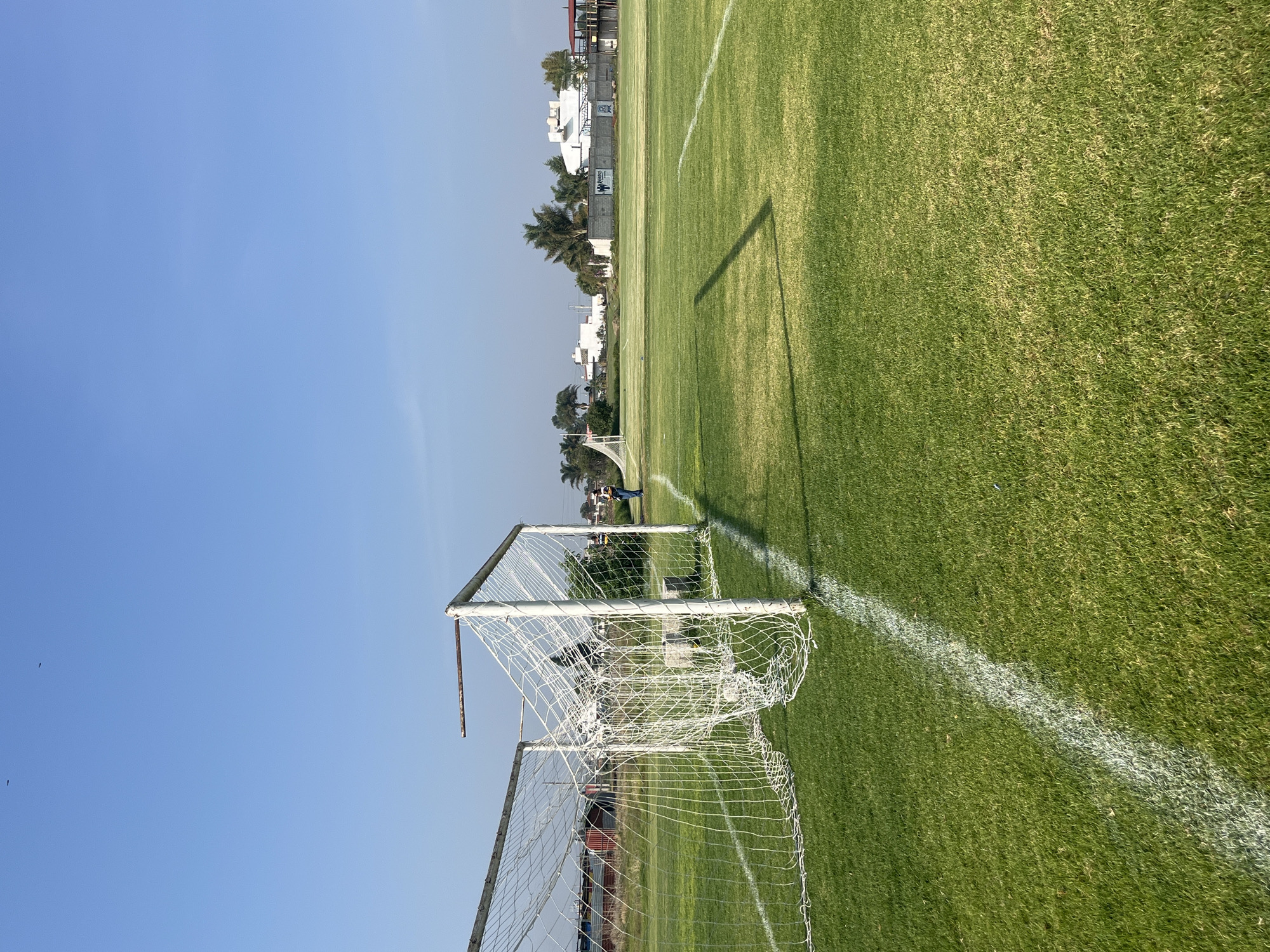 The image shows a soccer field with a goal in the foreground. The field is green with white lines marking the boundaries. In the background are buildings and trees. The sky is blue with no clouds. There is a person standing near the goal. The location is Unknown, Unknown.