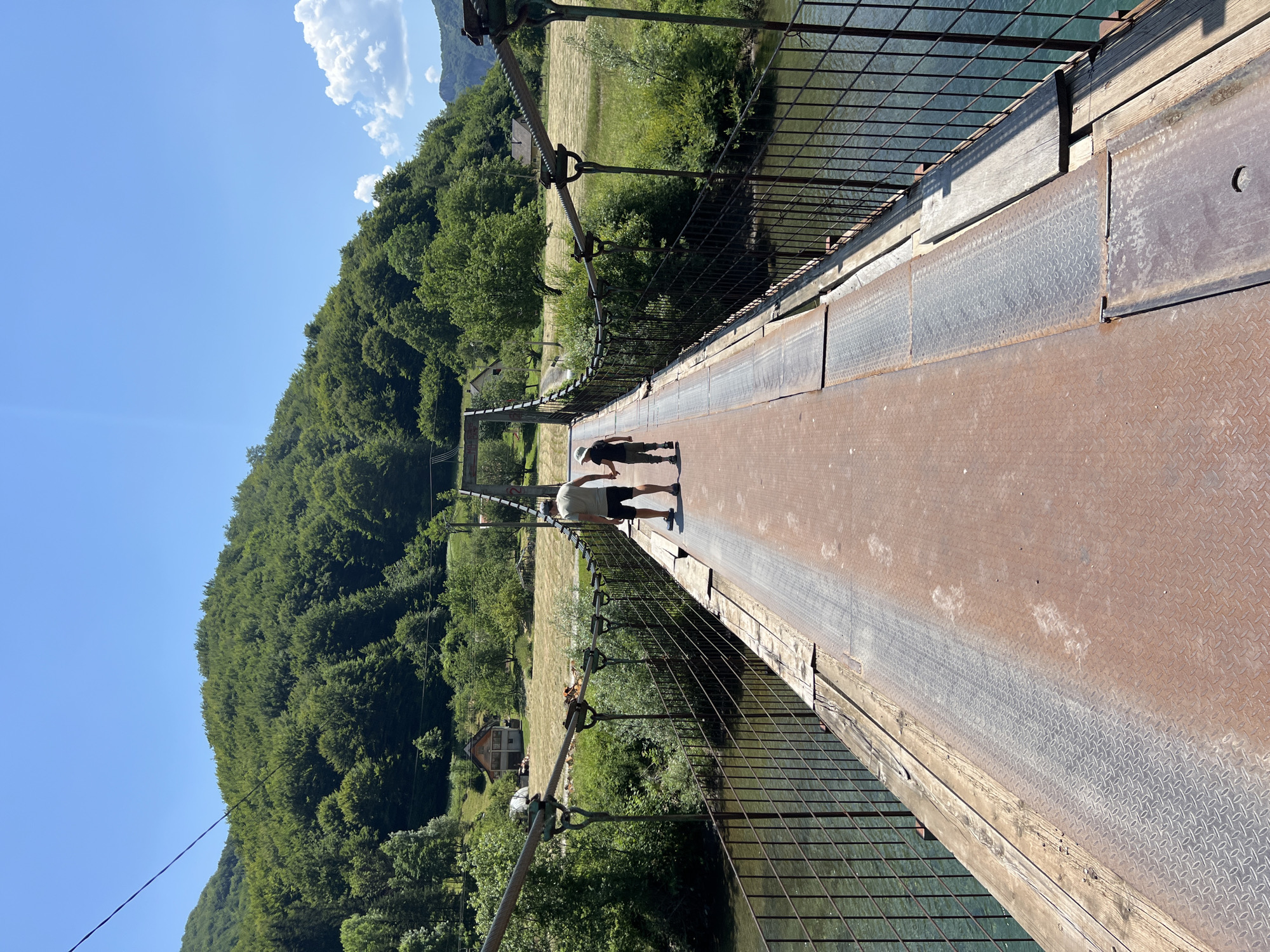 A man and a child are walking across a suspension bridge over a river. The bridge is made of metal and wood with a metal railing. The man is wearing a t-shirt and shorts, and the child is wearing a hat and shorts. They are walking away from the camera towards a wooded hill. The bridge is located in Moračko Trebaljevo, Montenegro. The environment is lush and green with trees and hills. The sky is clear and blue.