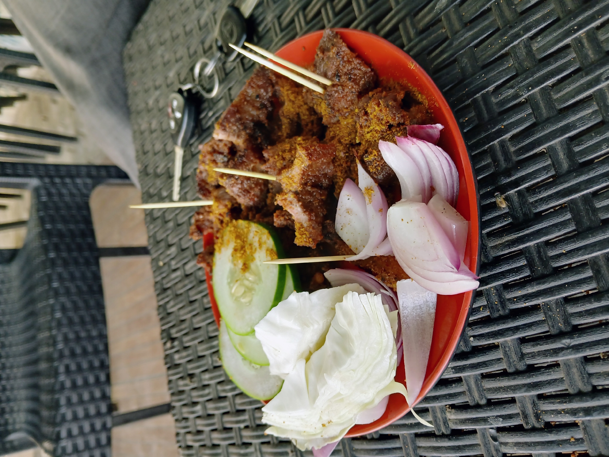 A red bowl containing meat skewers, sliced onions, cucumbers, and cabbage is sitting on a textured, woven surface. A set of keys is visible near the upper left corner of the bowl. The location is Barakin Lerawi, Nigeria.