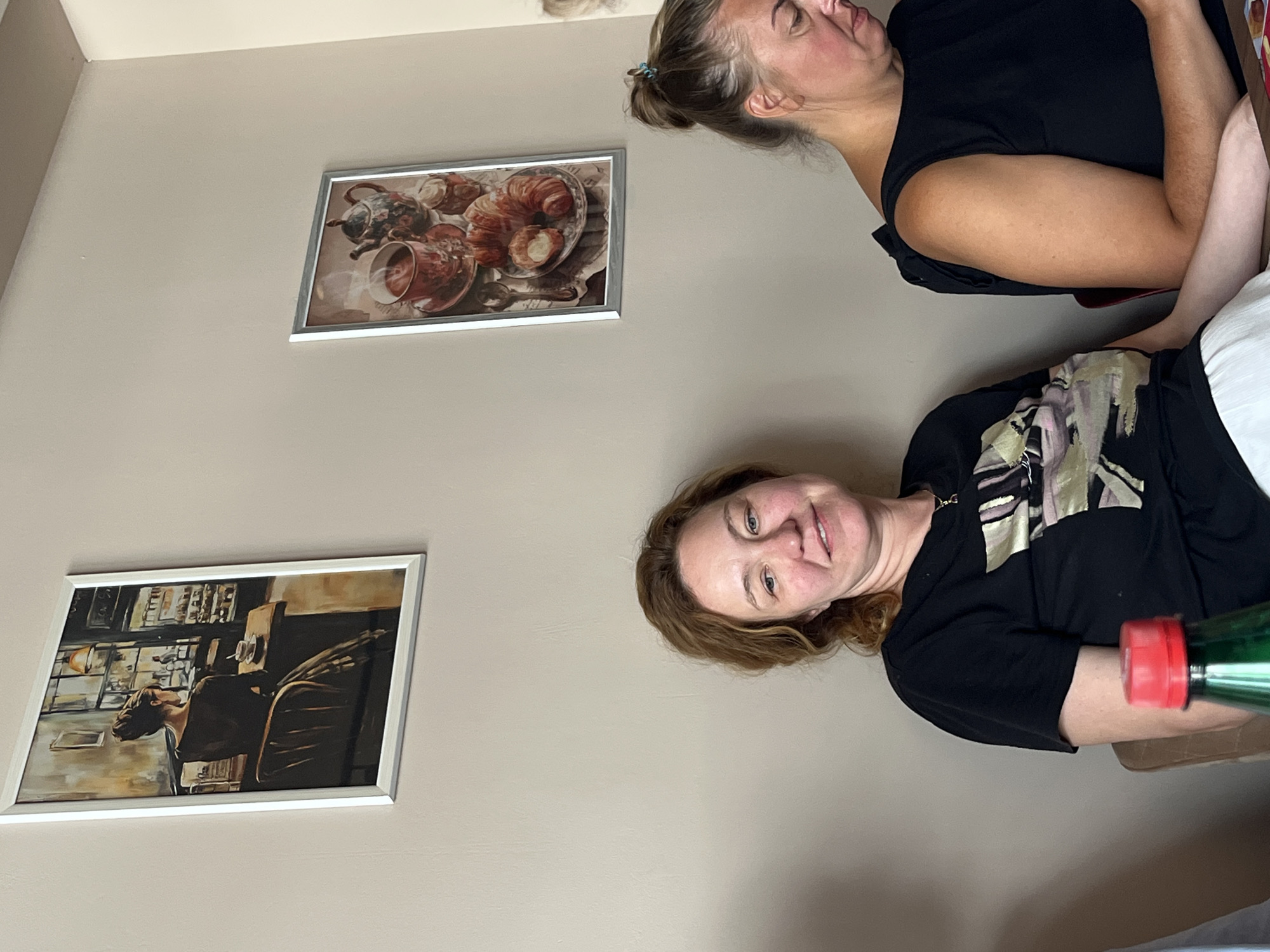 The photograph shows two women seated at tables in what appears to be a cafe in Donji Pobori, Montenegro. The woman on the left, with shoulder-length reddish-brown hair, is wearing a black t-shirt with a graphic design and white pants. She is looking slightly to her right, with a faint smile. The woman on the right, with her hair pulled back in a bun, is wearing a black sleeveless top. She is looking down at something on the table in front of her. Two framed pictures hang on the wall behind them. The picture on the left depicts a woman sitting at a table in a cafe, seen from behind. The picture on the right shows a tea set, croissants, and a bowl of sugar. A green bottle with a red cap is partially visible in the foreground on the left. The lighting suggests it is daytime, and the environment is indoors.