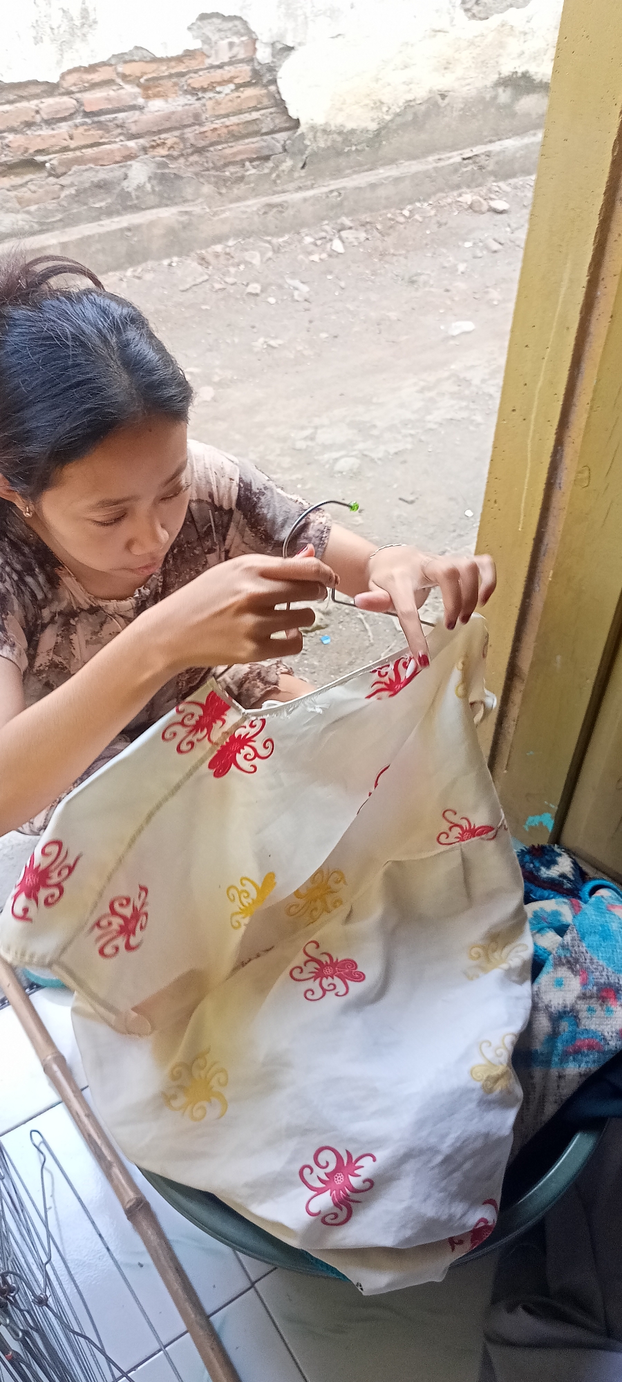 A young woman is sitting outdoors near a wall in Gempolklutuk, Indonesia, and is holding a metal hanger over a white fabric with red and yellow octopus-like patterns. Her hair is pulled back in a ponytail, and she is wearing a brown and white patterned shirt. The background shows a weathered brick wall and a yellow door frame. Several metal objects, possibly other hangers or tools, are visible on the ground to the left of the woman. To the right, a blue and patterned textile is partially visible. The ground appears to be paved with tiles and dirt.