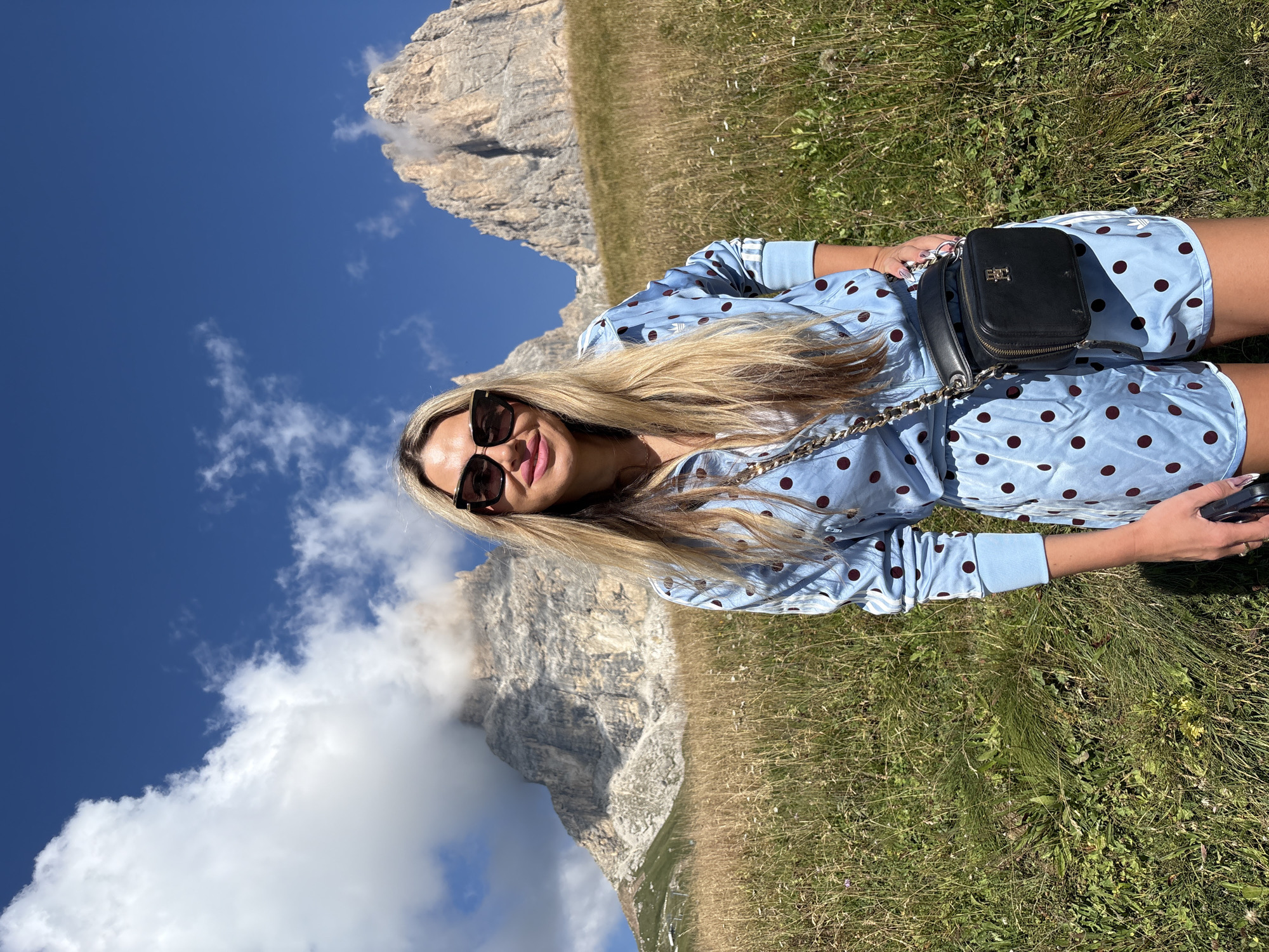 A woman stands in a grassy, mountainous area with a light blue, polka-dotted tracksuit. She is wearing large, dark sunglasses and a black crossbody bag with a gold chain strap. Behind her, a large, rocky mountain peak is visible, partially obscured by clouds. The sky is blue with scattered white clouds. The location is the Sella Pass, near Canazei, Italy.