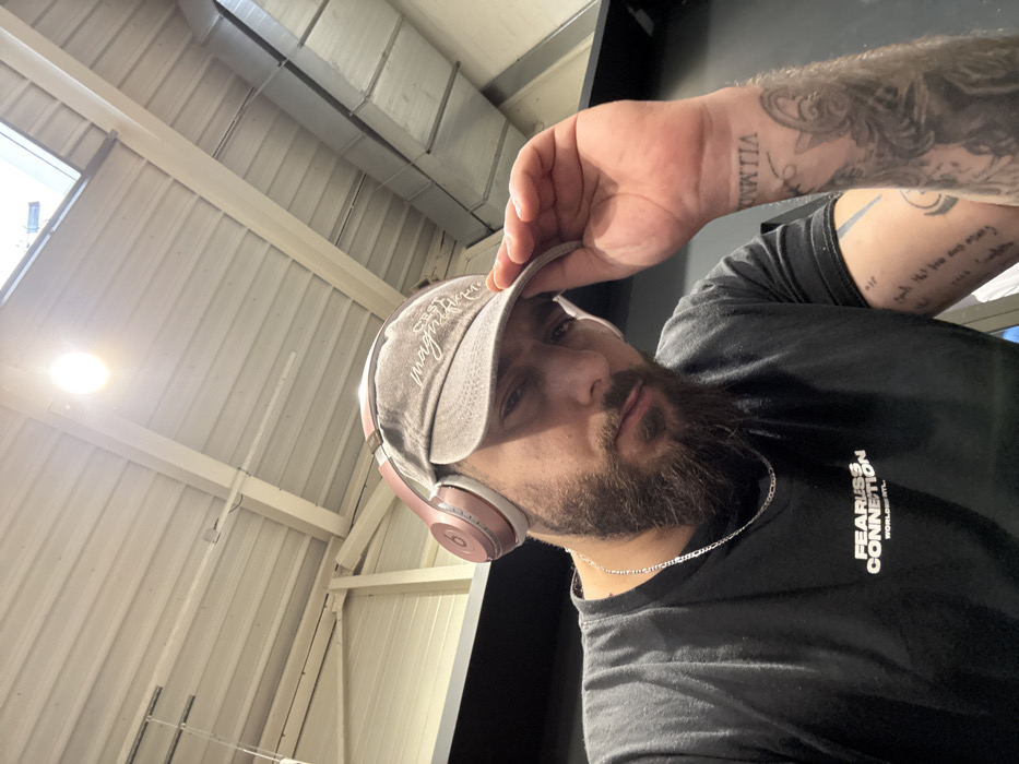 An adult male subject is depicted in a close-up, angled selfie within an indoor facility in Strasbourg, France. He possesses a dark beard and is wearing a black t-shirt with "FEARLESS CONNECTION WORLDWIDE ATL." printed in white, a silver-colored chain, rose gold over-ear headphones, and a faded grey baseball cap with "C'EST MAGNIFIQUE" embroidered on it. His left hand is positioned on the brim of his cap. Visible tattoos include a large figurative design on his left forearm, along with script and what appears to be stylized Roman numerals "VIIMMIIA." Script tattoos are also visible on his right forearm. The background consists of a high, light-colored corrugated metal ceiling supported by beams, a visible skylight, a bright overhead light fixture, and exposed metallic air ducts, suggesting an industrial or commercial space like a gym or warehouse. The walls in the background are dark. The subject maintains direct eye contact with the camera.