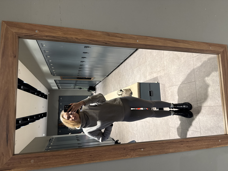 A person stands in front of a mirror taking a selfie with a phone. The reflection shows a locker room with rows of gray lockers and a bench along the wall. The person wears a gray turtleneck sweatshirt, gray leggings, and black boots. The mirror has a wooden frame. The lighting in the locker room is bright.