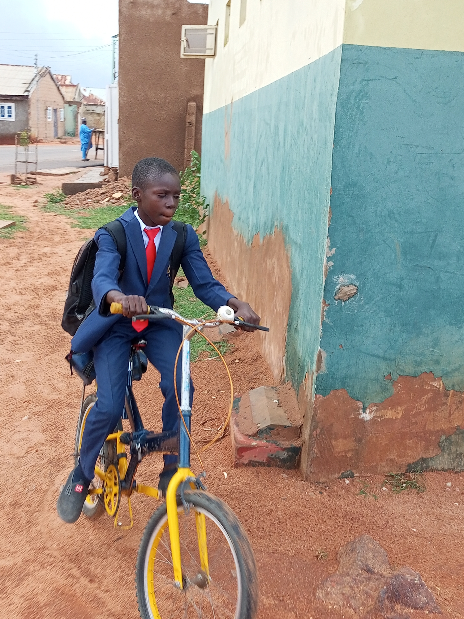 In Tumfure, Nigeria, a young boy wearing a navy blue school uniform with a red tie and a black backpack rides a bicycle. The bicycle is primarily blue with yellow accents on the wheels and pedal crank. The boy is looking to his right, with his hands on the handlebars. The environment is dusty and consists of a dirt path next to a building with a blue and beige facade. In the background, more buildings and a person in a blue garment are visible, suggesting a residential area.
