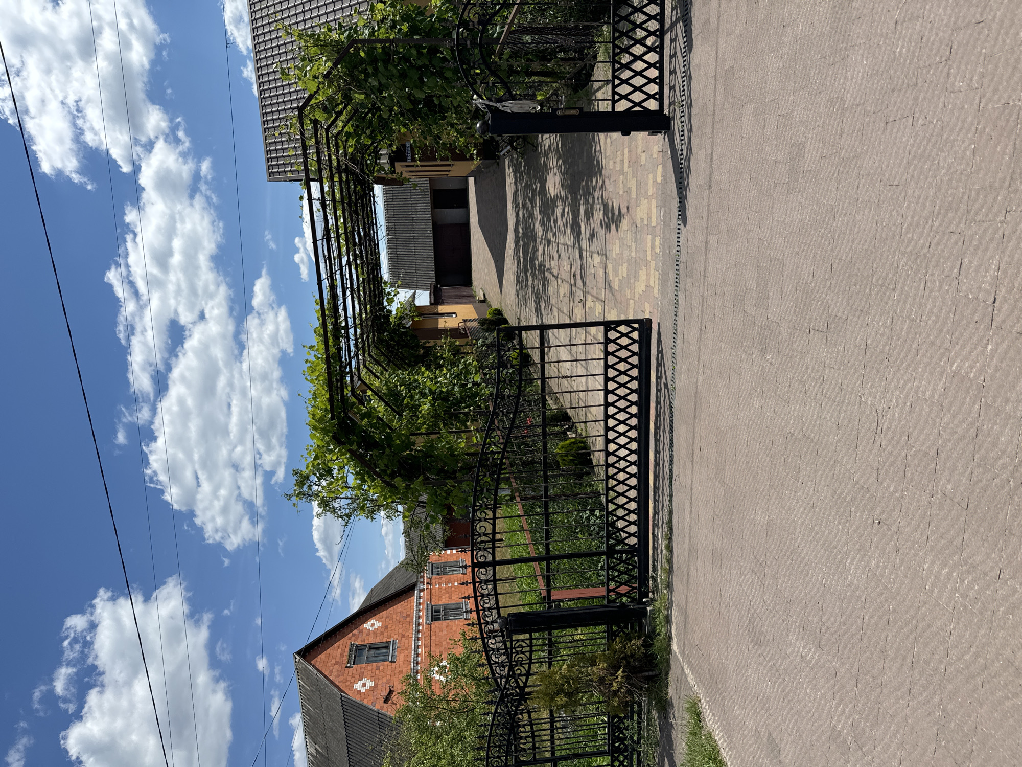 In Dovhe, Ukraine, a paved driveway leads to a property featuring a house and a garage. The house, constructed from red brick, has a dark-tiled roof and white decorative elements. Adjacent to it, a yellow building with a similar dark roof is visible. The driveway is partially covered by a metal pergola with climbing plants, casting dappled shadows on the ground. A black metal gate, partially open, guards the entrance to the property. The scene is set against a bright blue sky dotted with fluffy white clouds.