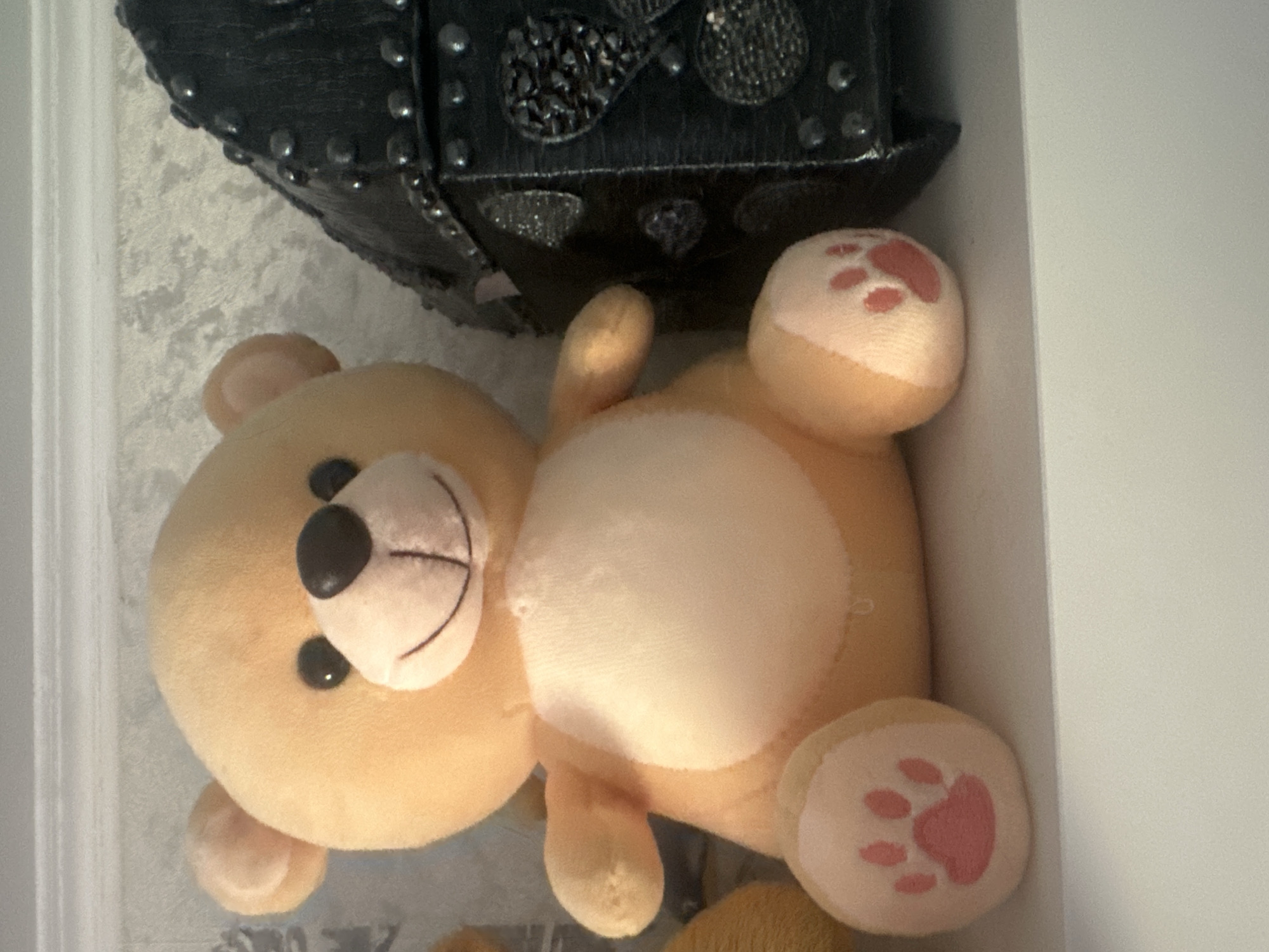 A light orange teddy bear sits on a white shelf. The bear is facing forward, with its paws on the shelf. The bear has black eyes and a black nose. The bear's belly is a lighter shade of orange. To the right of the bear is a black, spiked object. The background is a light grey textured wallpaper. The image was taken in Maladzyechna, Belarus.