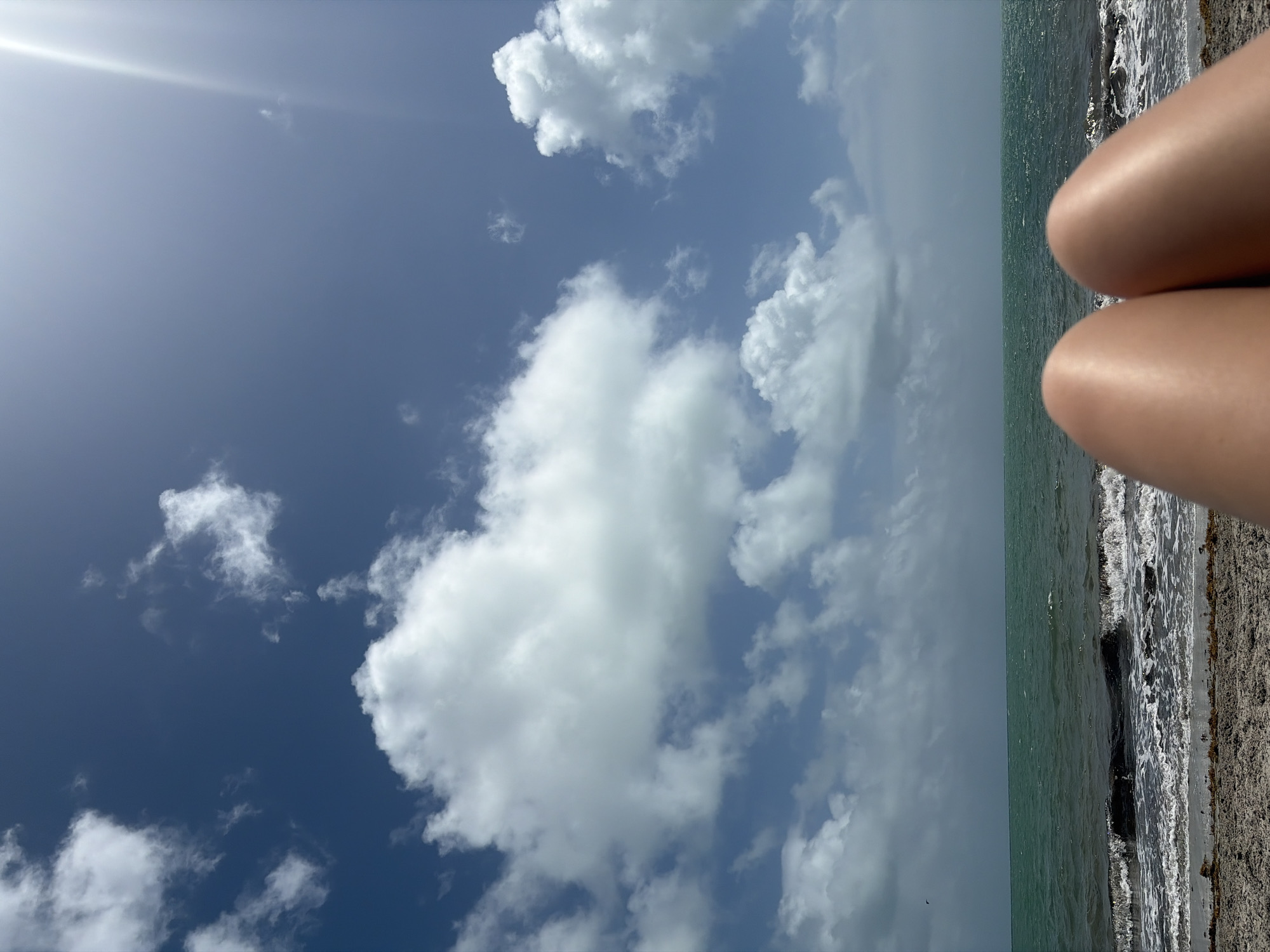 The image shows a scene at a beach in Key Biscayne, United States of America. The view is oriented towards the sky, which is predominantly blue with scattered white clouds and a visible sun glare. Part of the ocean and beach are visible, with waves crashing onto the shore. In the right edge of the frame, there are a pair of legs.
