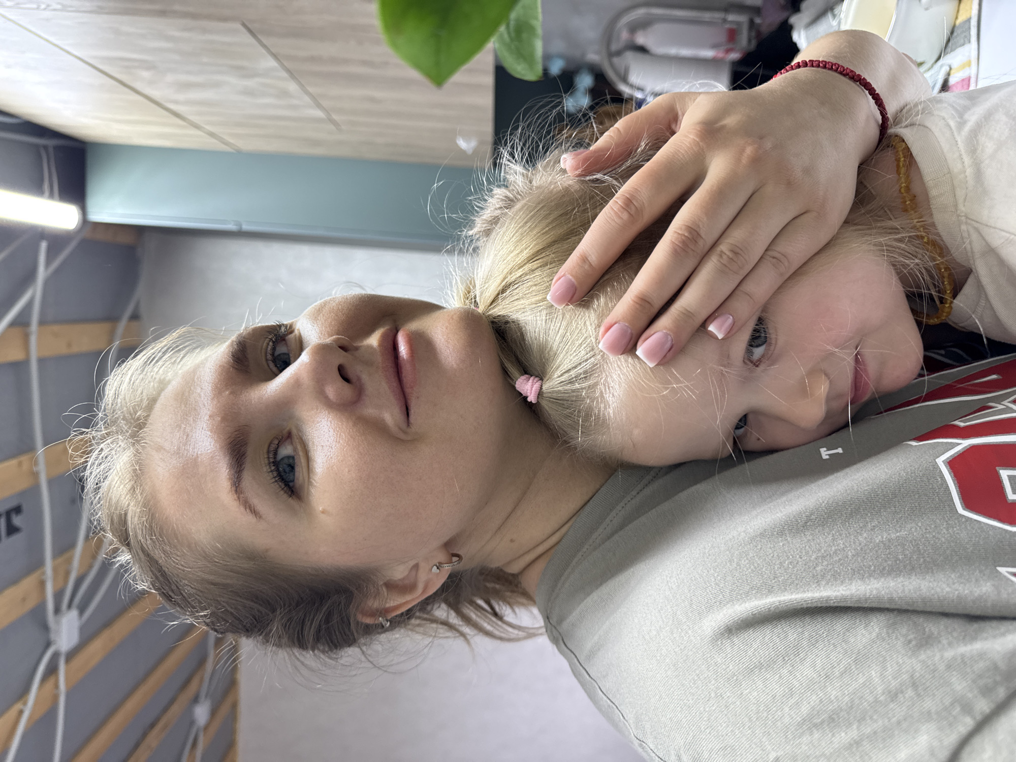 A woman and a child are lying down in Ферма, Russia. The woman is wearing a gray shirt and earrings, and she has blonde hair. The child is lying on the woman's chest, and the woman is holding the child's head. The child has blonde hair tied in a ponytail with a pink hair tie. The child is wearing a white shirt and has a red bracelet on her wrist. There is a plant in the background and a wall with wooden slats.