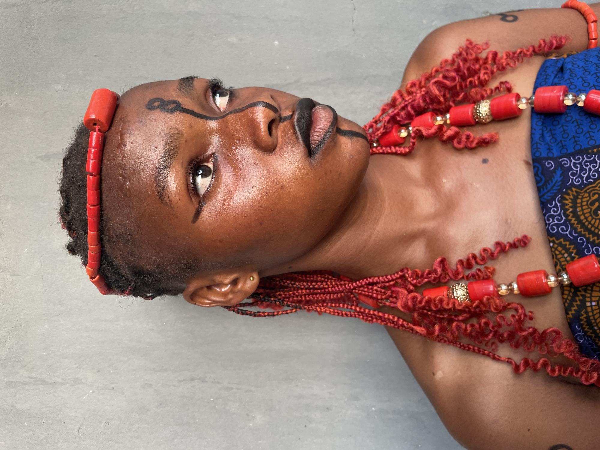 A young woman, likely in Badagry, Nigeria, is depicted lying on a neutral grey surface, looking upwards. Her dark skin is adorned with bold black body paint, featuring prominent linear designs around her eyes and across her lips, as well as a symbolic figure-eight or infinity-like marking on her forehead.
She wears vibrant red beaded jewelry, including a simple red beaded headband and elaborate strands of red beads, some interspersed with gold-colored cylindrical and spherical elements, draped around her neck and chest. Her hair appears short and dark, with decorative red curly braids or extensions interwoven with the beaded adornments. A blue fabric patterned with white and yellow motifs, possibly a traditional wrapper or garment, is visible on her right side. The lighting is bright and even, suggesting a daytime setting. Her expression is calm and contemplative. No discernible text is visible in the image.