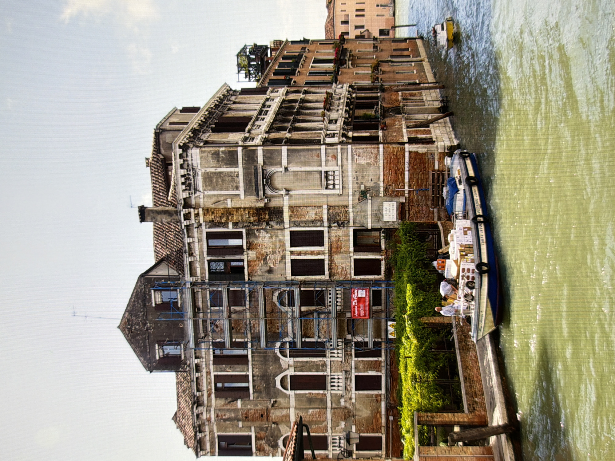 The image depicts a scenic canal view in Venice, Italy, not Kyiv, Ukraine. A large, multi-story building with a weathered facade and a red-tiled roof dominates the left side of the frame. Scaffolding is erected on the side of the building. In the foreground, a canal with greenish-yellow water flows. A blue and white boat is docked along the canal, carrying several boxes and attended by two individuals. Further down the canal, a small yellow boat is visible. The sky is overcast.