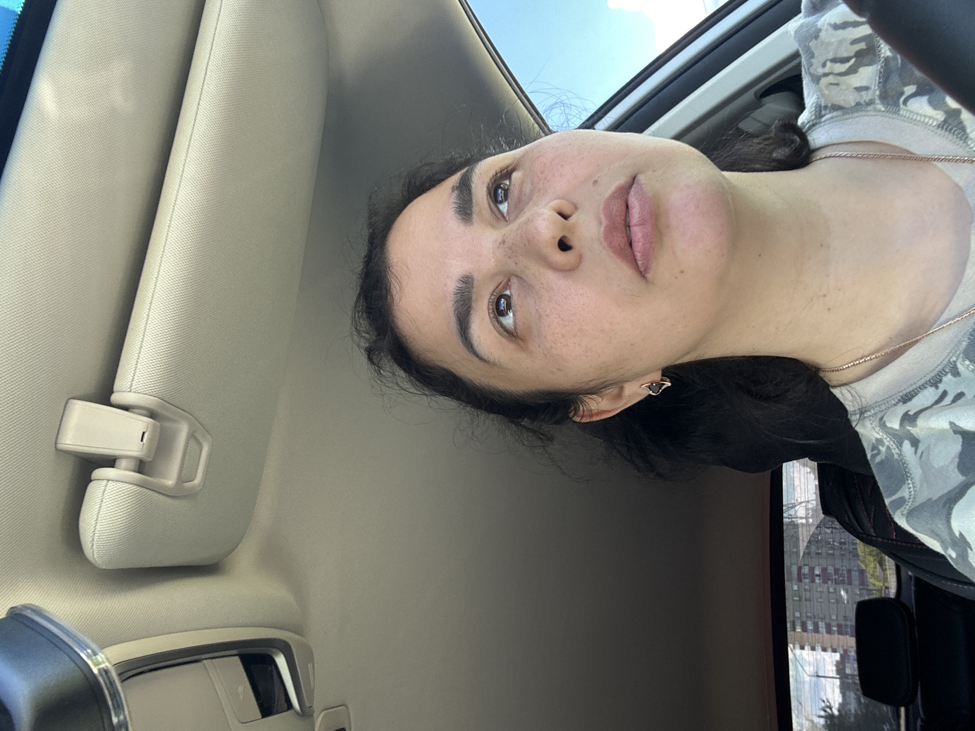 A woman with dark hair and freckled skin is pictured from inside a car, with the sun visor visible. Her shirt appears to be gray camouflage. Outside the car window, tall buildings of an unknown city are seen.