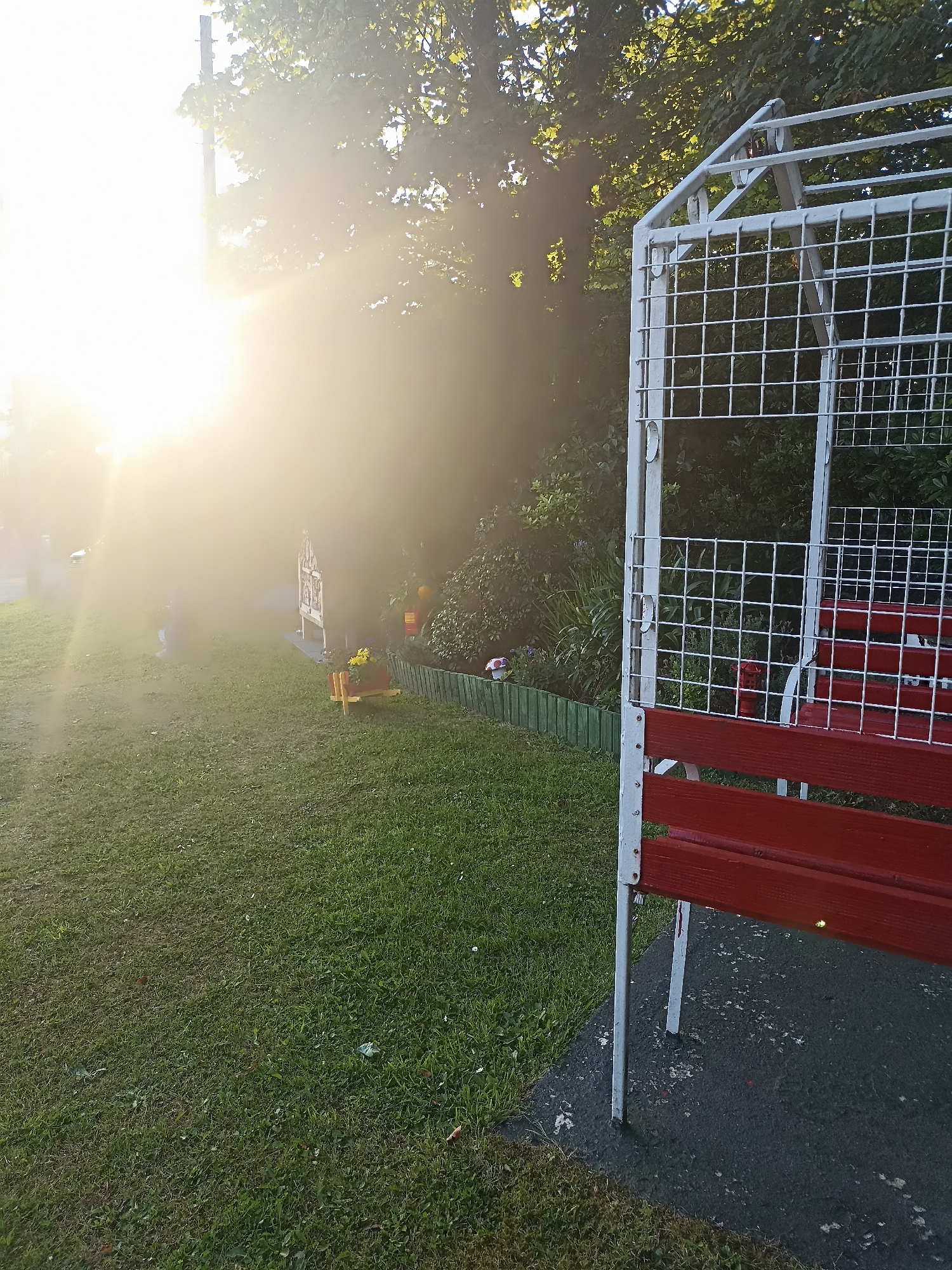 The image depicts a sunny outdoor scene, likely a garden or park area, with a white and red metal bench prominently featured in the foreground. The bench has red wooden slats and a white metal frame with a grid pattern. To the left of the bench, a patch of green grass is visible, leading towards a brighter area with trees and a power pole, suggesting a strong light source, possibly the sun, is present. In the background, there are lush green bushes, a small wooden planter with yellow flowers, and a green fence. The ground around the bench appears to be a dark, possibly asphalted area. The overall impression is that of a peaceful, well-maintained outdoor space. The specific location in Annagry, Ireland, cannot be determined from the image alone.