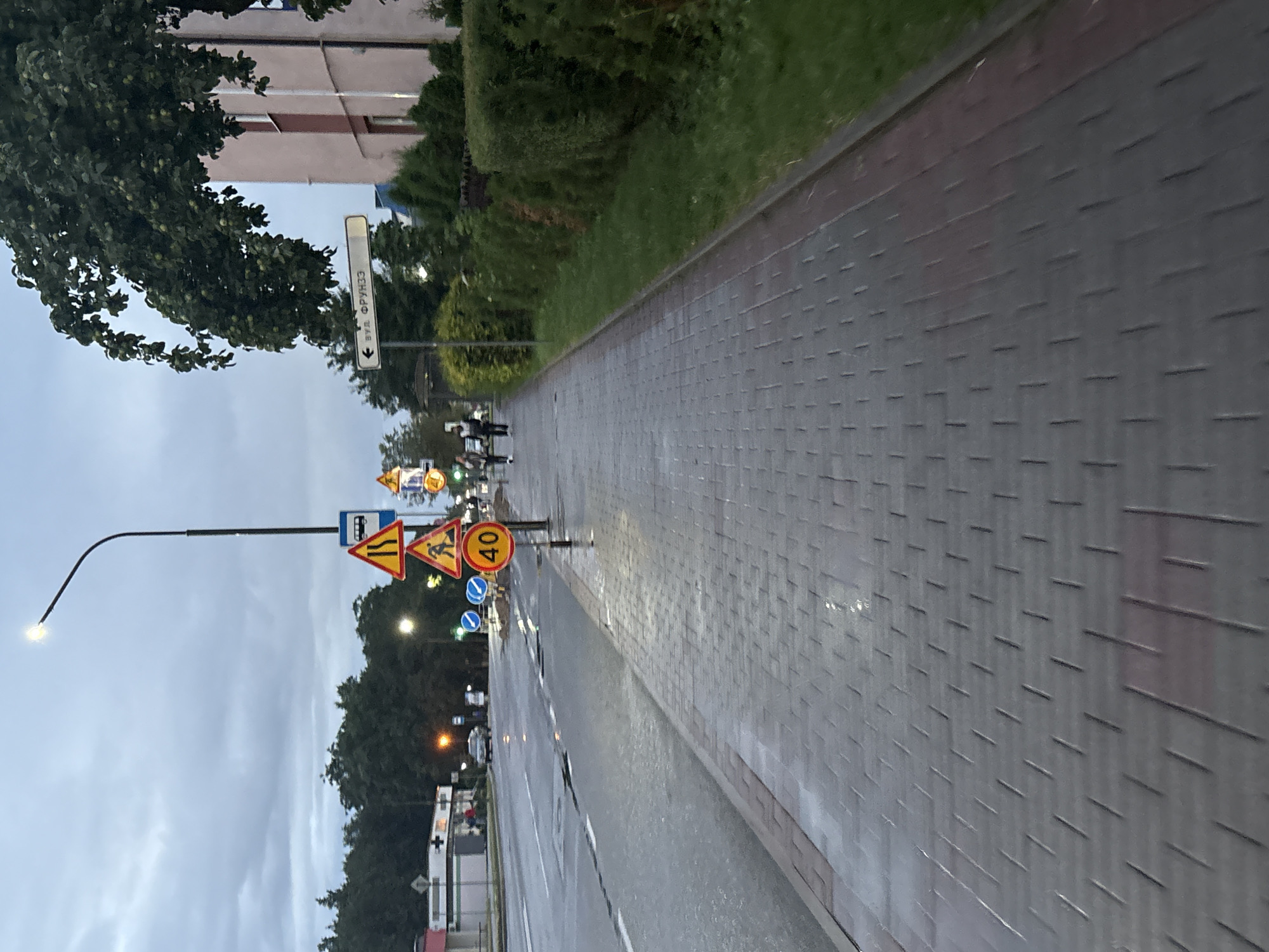 This media file captures an outdoor scene on a cloudy day, likely in the late afternoon or early evening, given the dim lighting and the illuminated streetlamp. The setting appears to be a street with a paved sidewalk on the right and a road on the left. The sidewalk is made of grey rectangular pavers laid in a repeating pattern, with a reddish-brown border. Adjacent to the sidewalk is a strip of green grass and then manicured bushes and trees, forming a hedge-like barrier. Beyond the greenery, a multi-story building with a light-colored facade is visible.
Several traffic signs are prominent. A street sign with Cyrillic text, reading "ВУЛ. ФРУНЗЕ", is mounted on a pole, indicating a street name. Other signs include a bus stop sign, triangular warning signs depicting road conditions such as merging lanes and pedestrian crossings, and a circular sign indicating a speed limit of 40. There are also two circular blue signs with white arrows.
In the distance, on the sidewalk, a few figures appear to be walking, suggesting pedestrian activity. A streetlamp with a curved arm is illuminated, casting a warm light. The road surface appears wet, possibly from recent rain, with reflections visible. The overall mood is calm and ordinary, depicting a typical urban environment under overcast skies. The location cues, such as the street sign, suggest the location is Lida, Belarus.