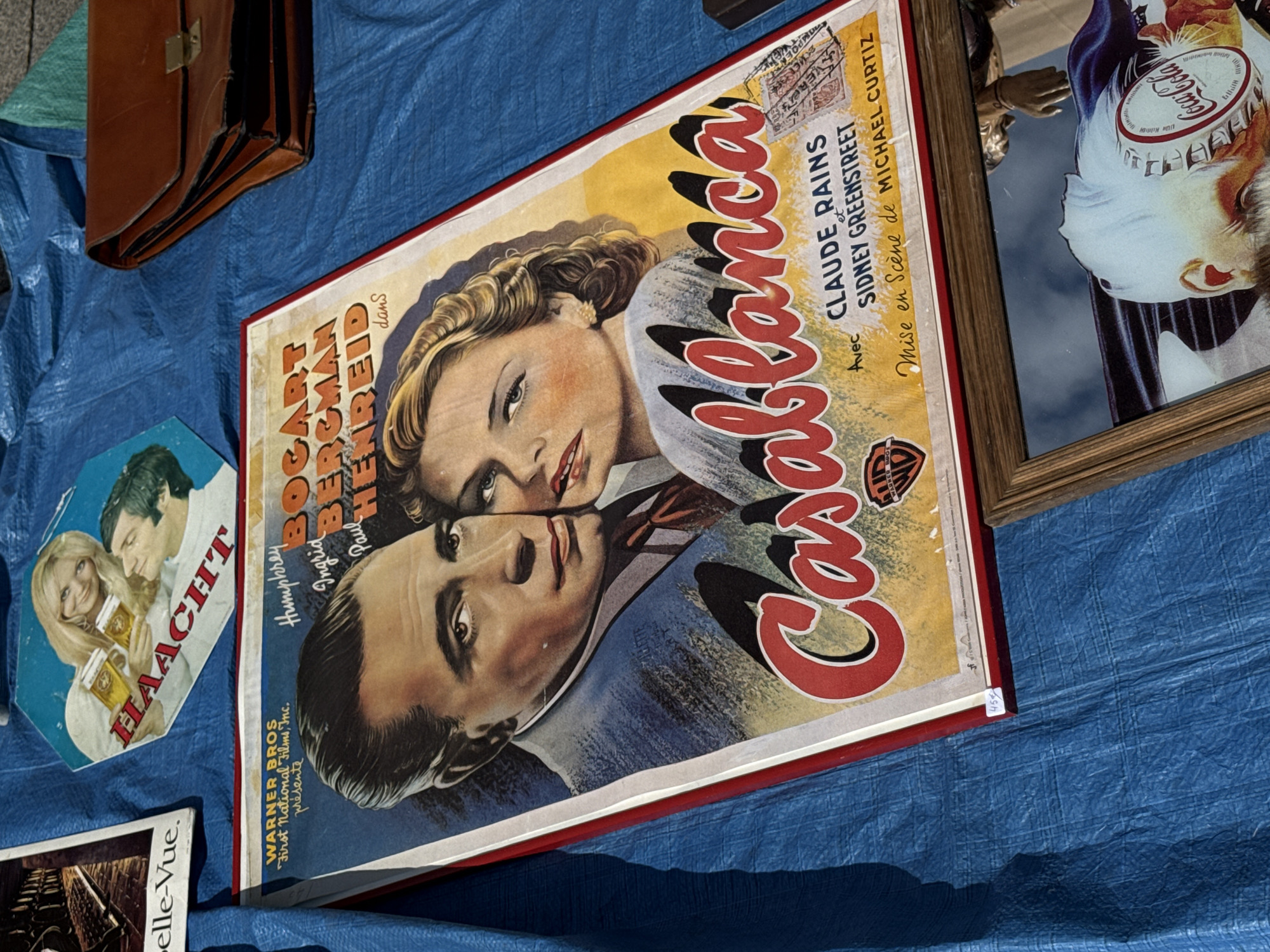 In Bruges, Belgium, on a blue tarp laid out for a market, rests a collection of vintage items. The most prominent is a large movie poster for "Casablanca," featuring portraits of Humphrey Bogart and Ingrid Bergman. The poster is framed by red and white borders. To the left of the poster, a smaller rectangular image shows what appears to be a street scene, with text that partially reads "belle-Vue." Above the "Casablanca" poster, a smaller triangular promotional item depicts a blonde woman and a man raising glasses of beer, with the word "HAACHT" printed in bold letters. To the right of the "Casablanca" poster, a brown leather briefcase is visible. In the foreground, a framed object, possibly a mirror or a picture, reflects an image of a person's head wearing a Coca-Cola cap. The overall impression is of items for sale at an outdoor market.