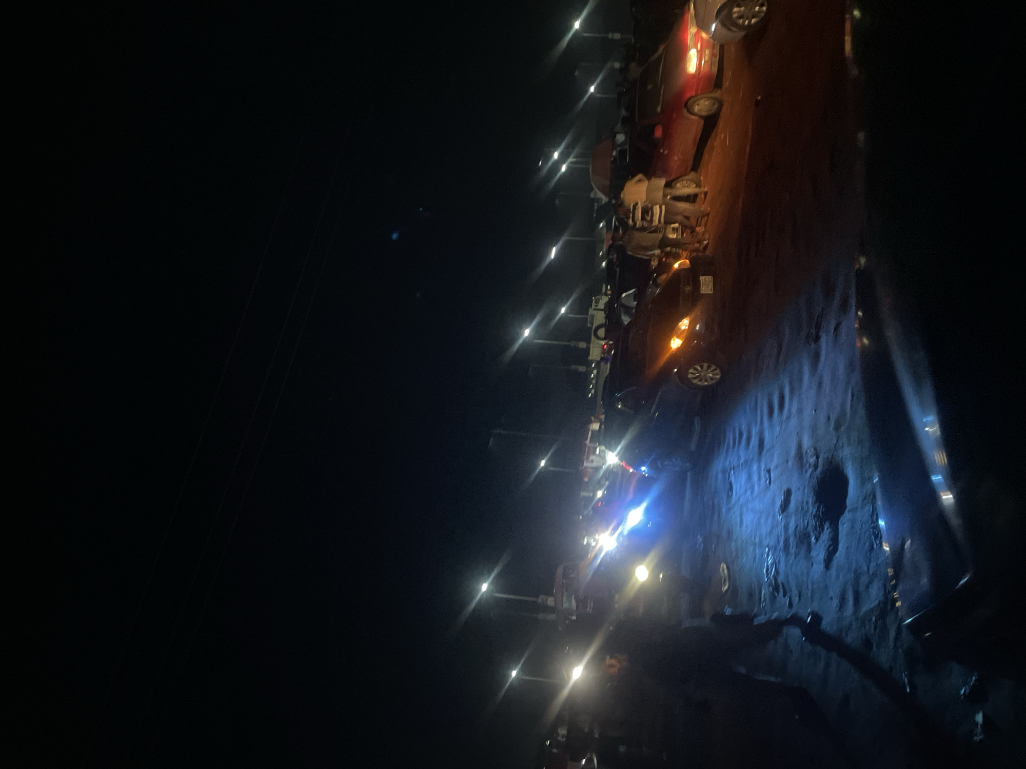 The image captures a nighttime scene on a dark, unpaved road in Sauka, Nigeria. The road surface is uneven and appears wet or muddy in places, reflecting the bright lights from passing vehicles and streetlights. A line of parked vehicles, including cars and what appear to be motorcycles or scooters, is visible along the side of the road. Some vehicles have their headlights on, illuminating the surrounding darkness.
The activity appears to be people gathered or interacting along the roadside. The presence of multiple vehicles suggests a potentially busy area or an event. The lighting is characterized by distinct, sharp streaks of light, indicative of a long exposure photograph or a camera struggling with low light conditions and bright light sources. The overall atmosphere is one of a typical nighttime street scene in a Nigerian locale, with the focus on the interplay of artificial lights and the dark environment. There are no discernible people clearly visible as distinct subjects, but their presence is implied by the parked vehicles and the general activity. The setting is clearly outdoors, on a road, under the cover of night.