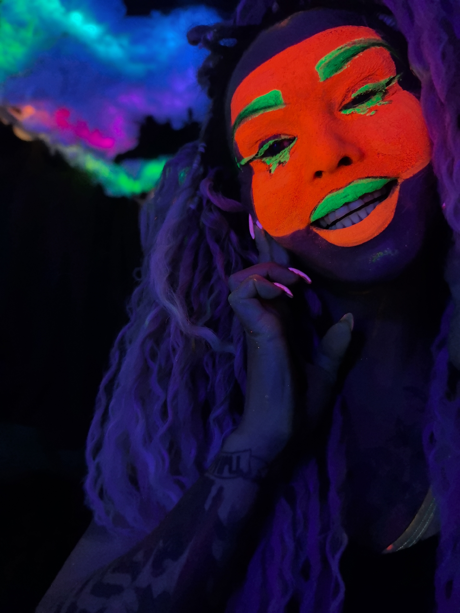 This image features a person, likely a woman, with striking glow-in-the-dark makeup and hair, set in a dark environment illuminated by UV light. Her face is painted a vibrant neon orange, with bold neon green eyeliner, eyeshadow, and lipstick. Her lips are parted in a subtle smile, revealing her teeth. Her long, wavy hair is a bright, glowing purple, complementing the neon colors of her makeup.
The person is posing with her right hand raised to her chin, showcasing long, manicured nails that glow with a pinkish-purple hue. A tattoo with indistinct glowing symbols or text is visible on her right arm. In the upper left background, a colorful, cloud-like element glows with various neon shades of blue, green, pink, and purple, contributing to the surreal and energetic atmosphere.
The scene is dominated by darkness, typical of a blacklight setting, suggesting an indoor event such as a party, club, or artistic photoshoot. The focus is entirely on the person's elaborate and luminous appearance, which creates a visually captivating and artistic impression. No specific location cues are visible, and the time of day is irrelevant due to the artificial lighting conditions.