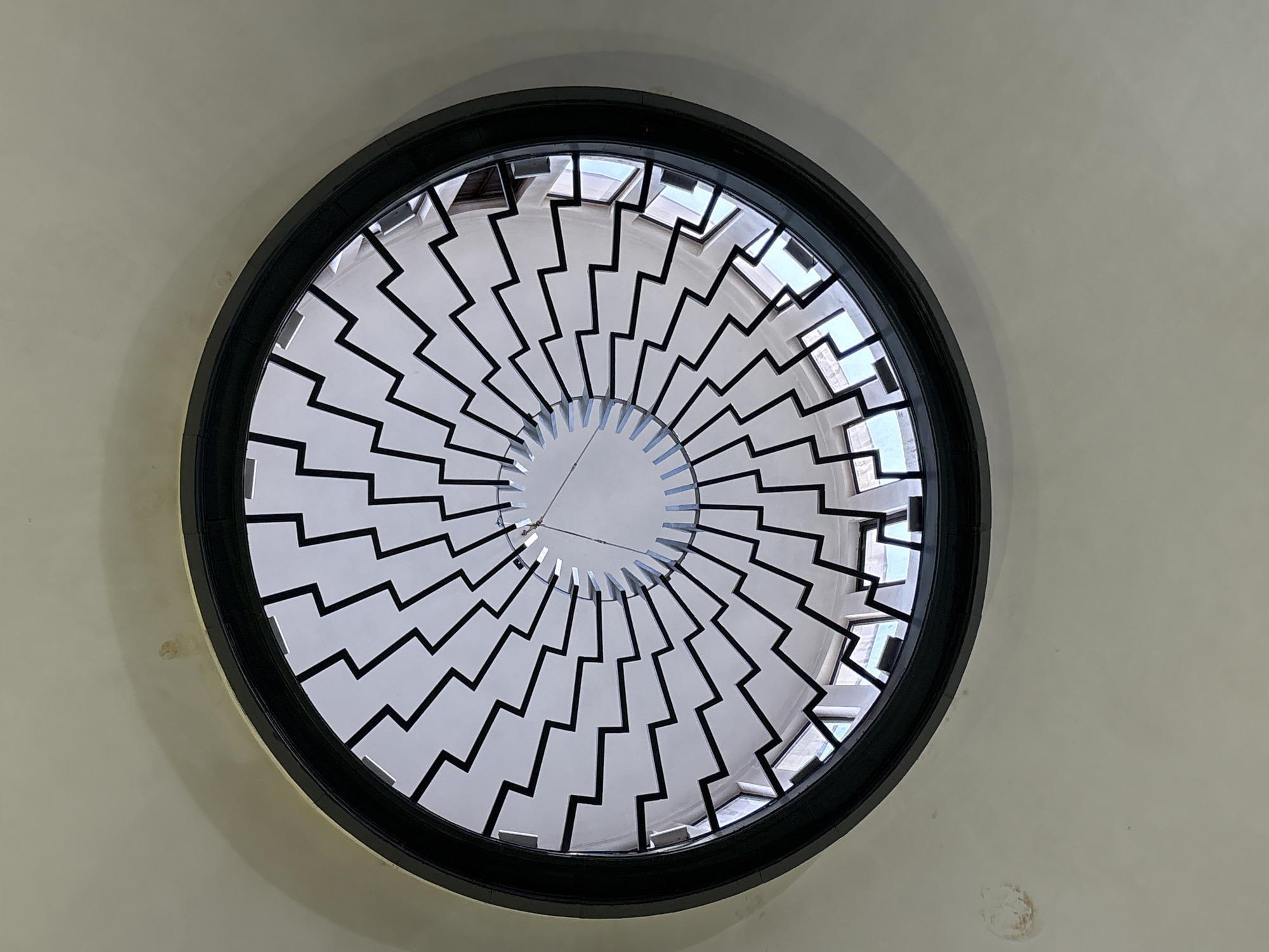 The image is a low-angle view of a circular skylight set into a light beige ceiling. The skylight has a thick, black frame. Within the frame is a decorative metal structure composed of evenly spaced, zigzag-shaped elements radiating from the center. The spaces between these zigzag elements allow light to pass through. Looking through the skylight, hints of other architectural structures are visible in the gaps. A thin wire or string extends across the center of the skylight. The overall impression suggests a modern architectural design element.
Based on architectural style, this image is plausibly located in Saint Petersburg, Russia. However, this cannot be conclusively confirmed from the image alone.
