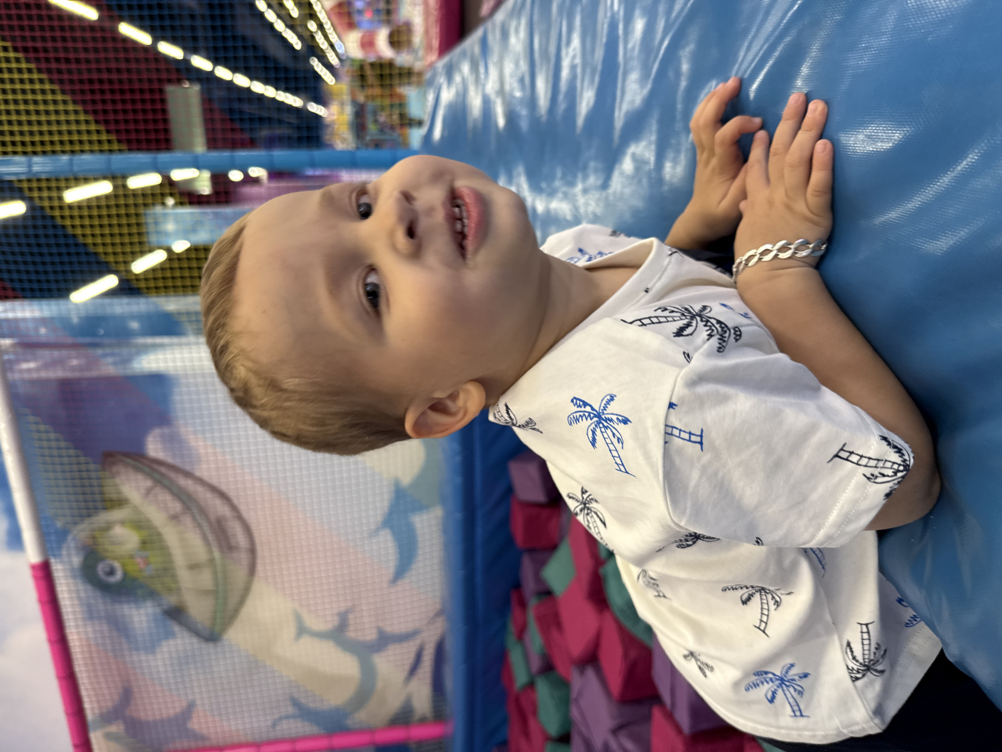 A young boy with blonde hair and blue eyes lies on a blue padded surface, looking towards the camera with a slight smile. He is wearing a white t-shirt with a pattern of blue palm trees. His left arm is bent at the elbow, and his left hand rests on the blue surface, adorned with a silver bracelet. In the background, there is a ball pit filled with purple, pink, and green foam cubes, and a colorful netted structure with a cartoon image of a green alien in a flying saucer. The scene appears to be inside a children's play area, possibly a children's entertainment center. The image is taken from a medium shot, with the boy as the primary subject. The lighting is bright, indicating an indoor artificial light source. The location is identified as Zhukovo, Russia, suggesting this is a local establishment.