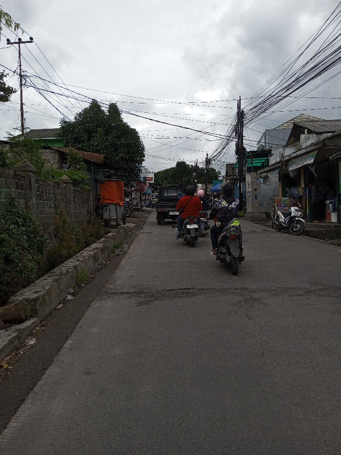 The image displays an overcast daytime street scene in the city of Melakudikadu, India. An asphalt road, exhibiting visible wear and cracks, occupies the foreground and recedes into the distance. On the left side, a concrete curb borders a shallow drainage ditch, behind which a low stone wall is partially covered by green vegetation. Further along the left, a stall-like structure with an orange tarp cover is present. On the right side, multiple low-rise buildings, appearing to be commercial establishments with storefronts, line the road. One building features a corrugated metal roof extension over its entrance, and a white scooter is parked on the sidewalk nearby. Overhead, numerous utility poles support a dense network of tangled electrical and communication wires that crisscross the sky. Vehicle traffic is observed moving away from the viewer. In the mid-ground, a black pickup truck is visible, followed by three motorcycles. The first motorcycle rider wears a red shirt and a dark helmet. The second motorcycle, positioned closer to the viewer, has a rider wearing a black and white patterned jacket with green accents and a dark helmet. A third rider with a light-colored helmet is partially visible between the two leading motorcycles.