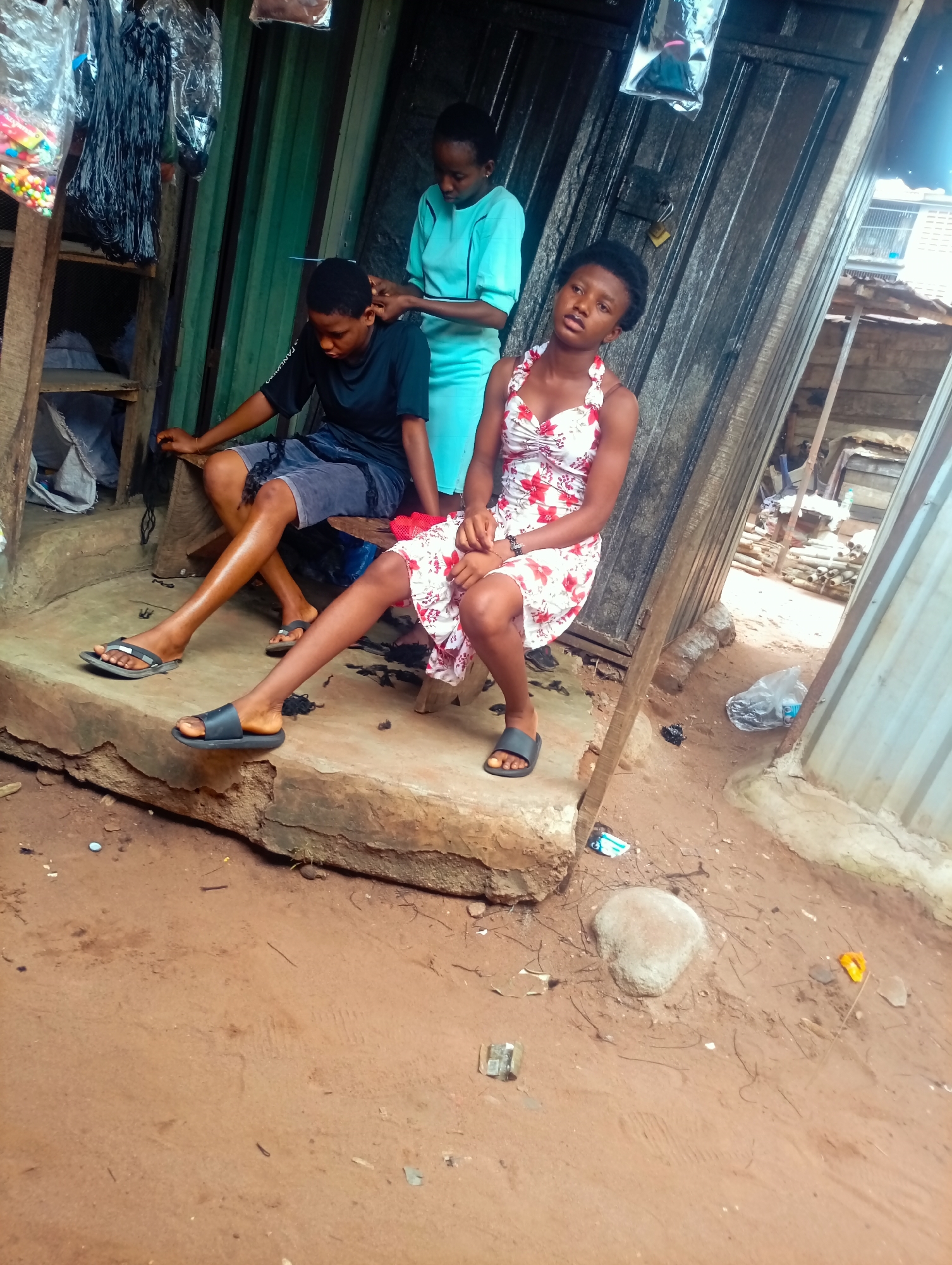 The image shows three young people in an outdoor setting, likely a market stall in Nkpor, Nigeria. In the foreground, a young woman with dark skin and short, styled hair is seated, looking towards the camera with a slightly weary or bored expression. She is wearing a white, floral-print dress with red flowers and thin straps. To her left, a young person with short hair, wearing a dark t-shirt and grey shorts, is also seated. They are looking down, and their hands are engaged with hair. Standing behind them, a young woman with dark skin and hair is braiding or styling the hair of the person in the t-shirt. She is wearing a light blue, short-sleeved dress.
The setting appears to be a simple, rustic stall. Behind the people, there are shelves with various items, including what looks like hair extensions or braiding materials and colorful beads. The structure appears to be made of wood and corrugated metal. The ground is sandy and reddish-brown, with scattered debris like small stones, leaves, and plastic. In the background, there are glimpses of other structures, suggesting a densely populated or commercial area. The lighting suggests it is daytime, with diffused light, possibly indicating an overcast sky or a shaded area. The overall atmosphere is informal and suggests a moment of everyday life, perhaps a hair salon or a personal grooming session taking place in a public or semi-public space.