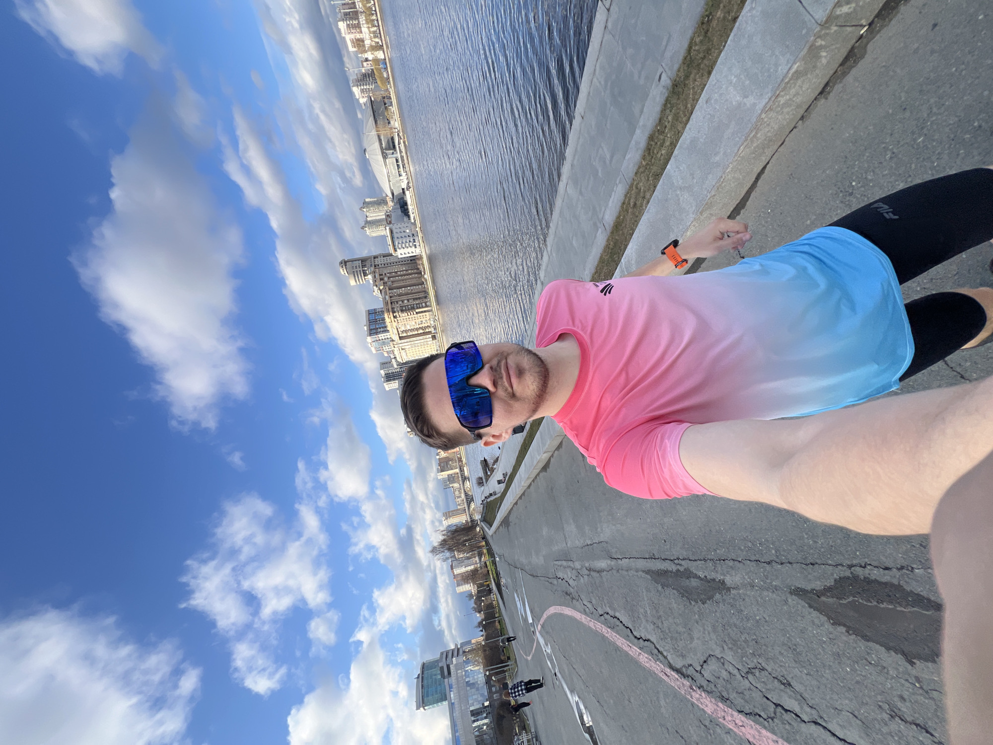 A man takes a selfie on a sunny day by the Iset River in Yekaterinburg, Russia. He is wearing sunglasses, a pink and blue ombre t-shirt, and black leggings. The background includes the river, a concrete embankment, city buildings, and a blue sky with scattered clouds. The ground is asphalt with a red line painted on it. Other people are visible in the background.