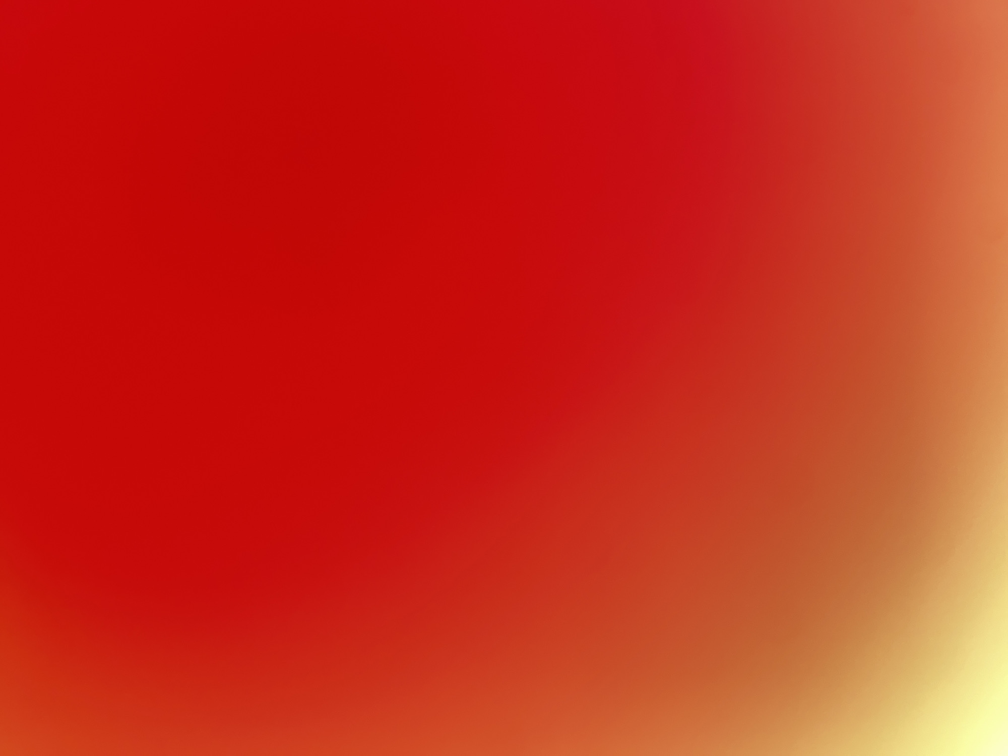 This is an abstract, out-of-focus image of a gradient of colors. The predominant colors are red and orange, transitioning to yellow at the bottom left corner. The image appears to be a digital artwork or a photograph of a light source or colored fabric, with soft, blurred edges. There are no discernible objects, people, or environmental features present. The location is unknown.