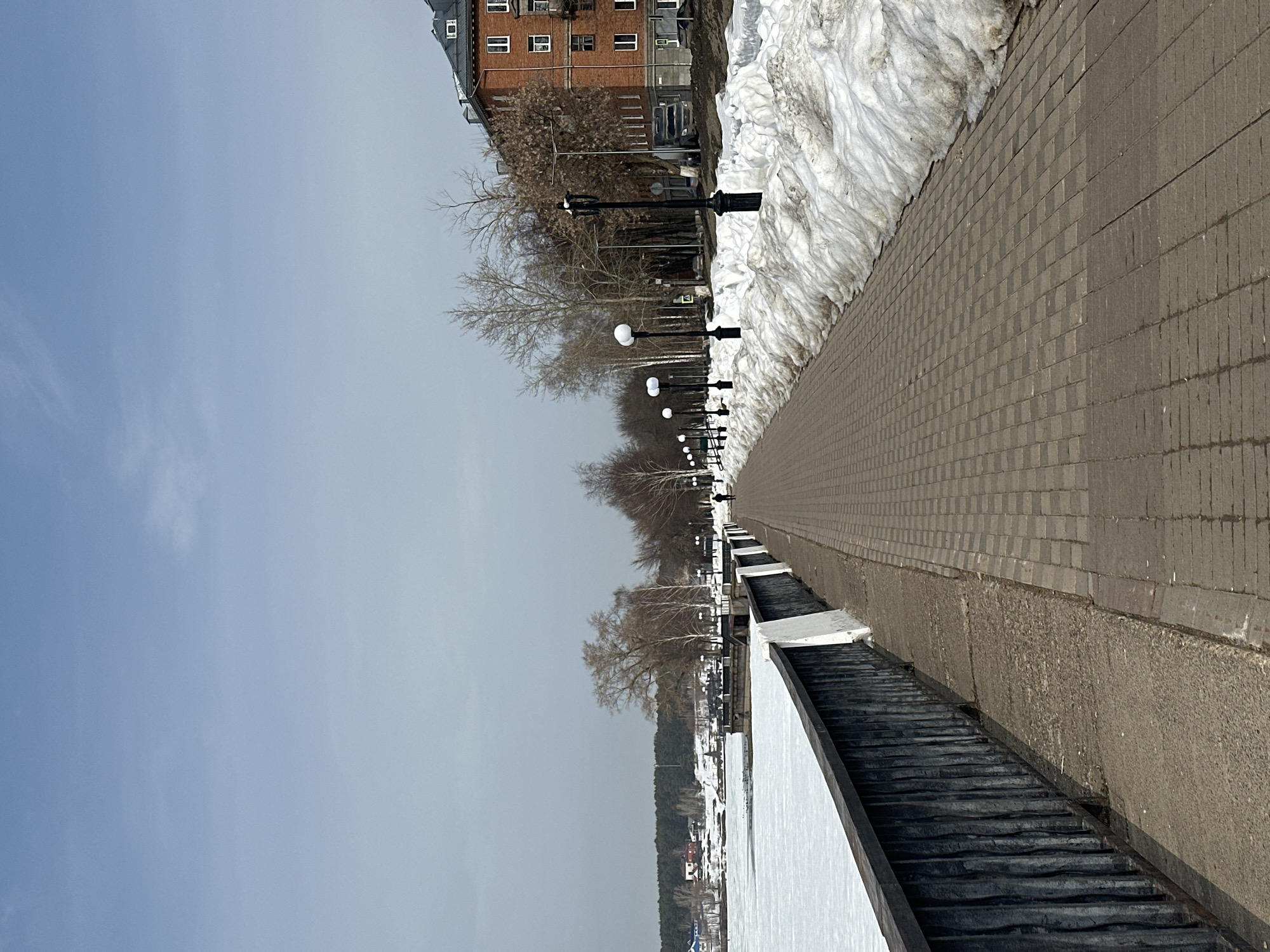 The image presents a wide, paved promenade alongside the Votkinsk Pond in Votkinsk, Russia, under a pale blue sky. The environment indicates a late winter or early spring setting.
On the left, a dark metal railing on a concrete base separates the promenade from the expansive, ice-covered surface of the Votkinsk Pond. This frozen expanse extends to a distant, snow-covered shoreline featuring sparse residential buildings with colored roofs and a dense coniferous forest. Closer to the far bank, dark patches on the ice suggest open water or thin ice, with indistinct forms possibly indicating waterfowl.
The promenade, constructed of rectangular grey pavers, extends in a straight line into the background. Along its right edge, a substantial, irregular pile of dirty, melting snow runs parallel to the walkway. A series of dark lampposts with spherical white globes lines the promenade, positioned between the paved surface and the snow. Behind the snow and lampposts, numerous bare deciduous trees line the embankment, leading to a multi-story, red brick building with multiple white-framed windows and a dark roof, partially visible on the far right.
A single, dark-clad individual is observed walking away from the camera in the distant section of the promenade.