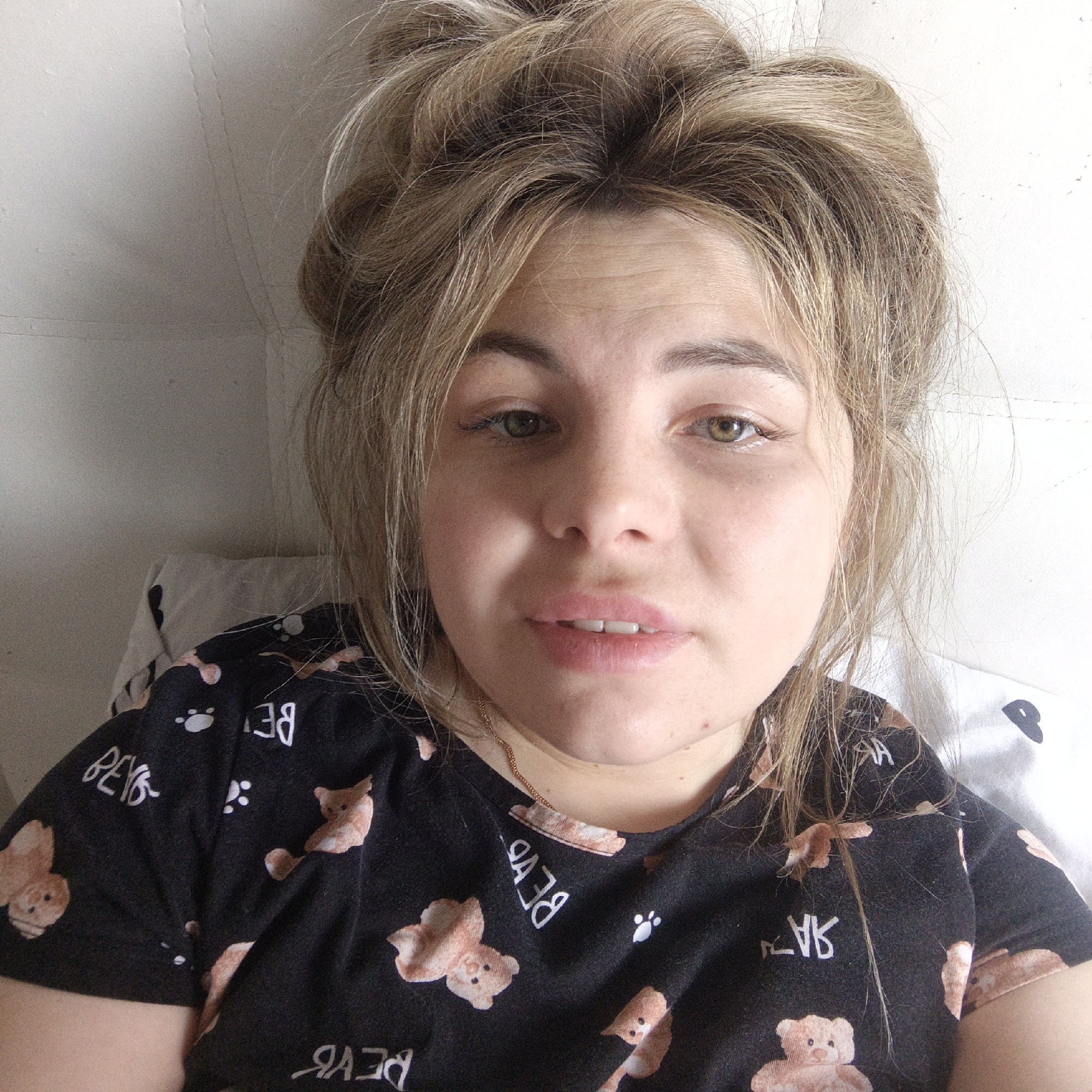 A close-up selfie shot shows a woman with light brown hair styled in a messy bun. Her skin tone is fair, and she has hazel eyes. She is wearing a black t-shirt with a teddy bear pattern and a thin gold necklace. The background appears to be a white pillow and a white wall. The location is Unknown, Unknown.