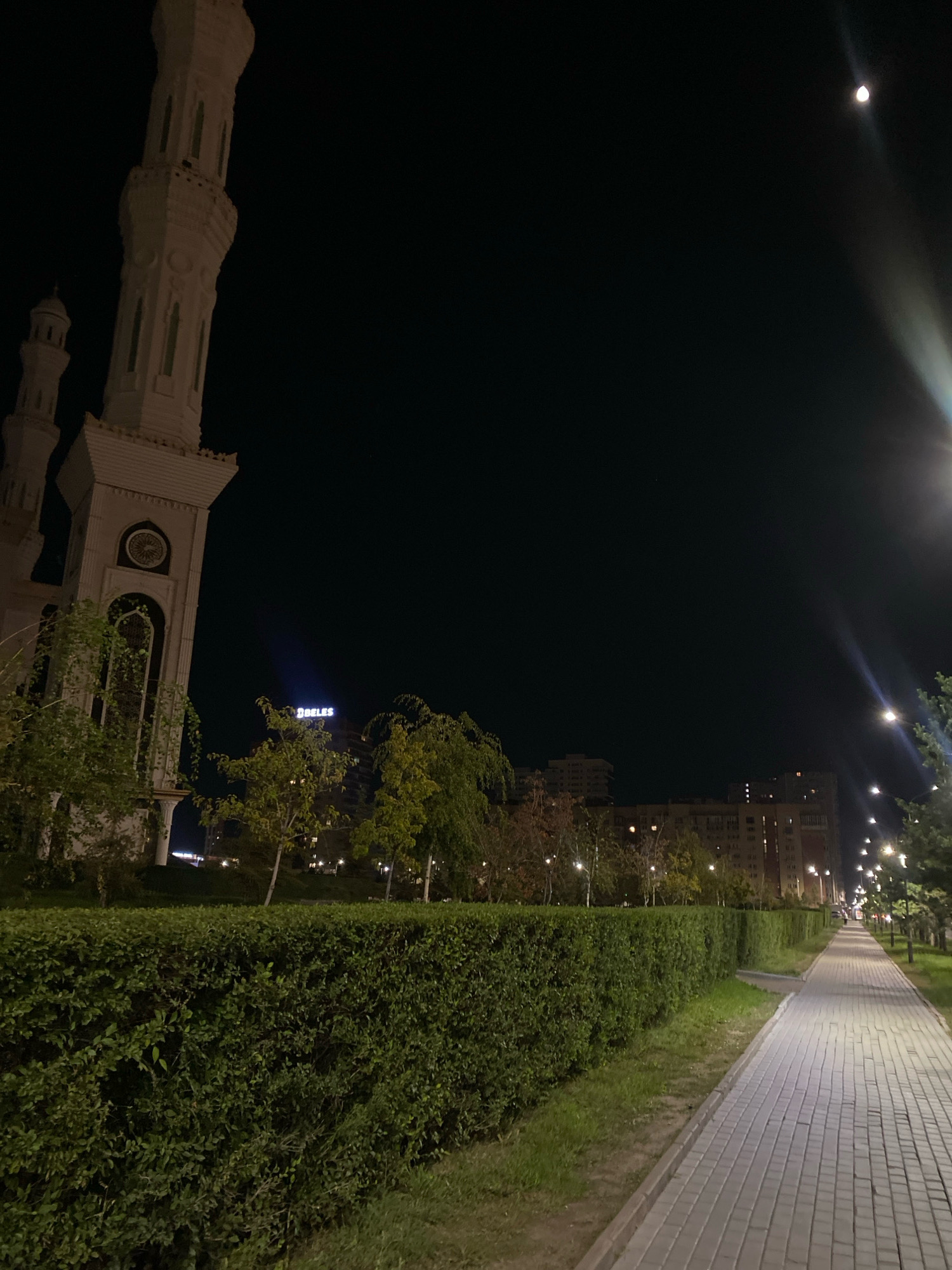 A nighttime view of a well-lit pedestrian walkway in Astana, Kazakhstan, alongside a manicured hedge. To the left, a section of a large, ornate minaret and part of a mosque are visible, bathed in artificial light. Beyond the hedge and trees, apartment buildings with illuminated windows form the cityscape. The moon is visible in the dark sky, and a street lamp casts a bright beam down the walkway. A sign with the word "ORDA" in blue illuminated letters is visible on one of the buildings.