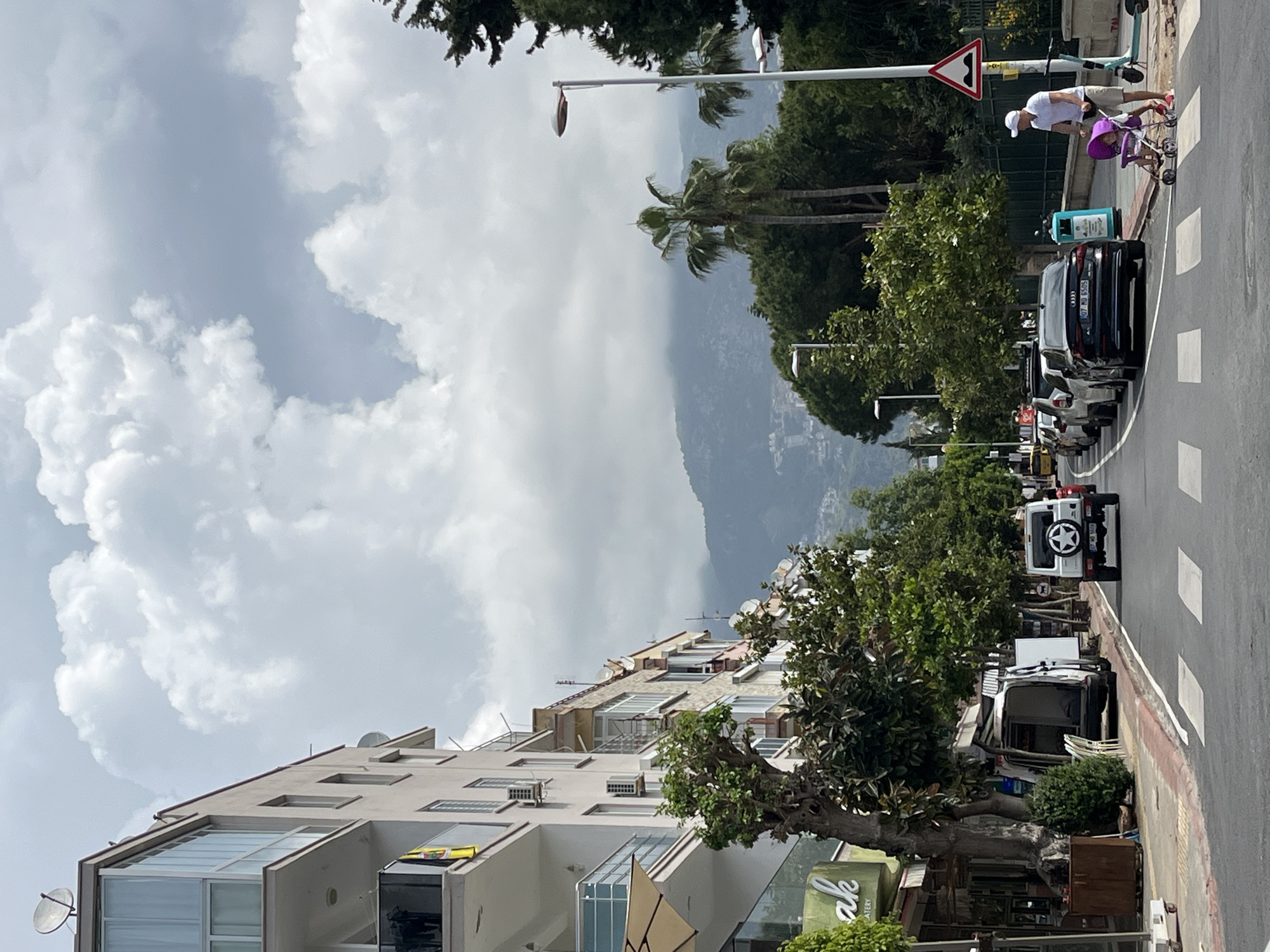 The image depicts a street in Alanya, Turkey, characterized by a mix of urban and natural elements. On the left, a multi-story building stands with apartments visible through glass-enclosed balconies. Satellite dishes are affixed to the building's exterior. Street-level commercial establishments are indicated by signage beneath the building.
A road runs alongside the building, lined with parked vehicles. A white SUV is prominently positioned, with its rear visible. Trees are interspersed along the sidewalk, adding greenery to the urban landscape.
In the background, a mountain range rises, partially obscured by clouds. The sky is overcast, with heavy cloud cover dominating the upper portion of the image. A palm tree is situated among the other trees along the roadside. A man is seen pushing a stroller along the sidewalk in the top right of the frame. A road sign is present nearby.