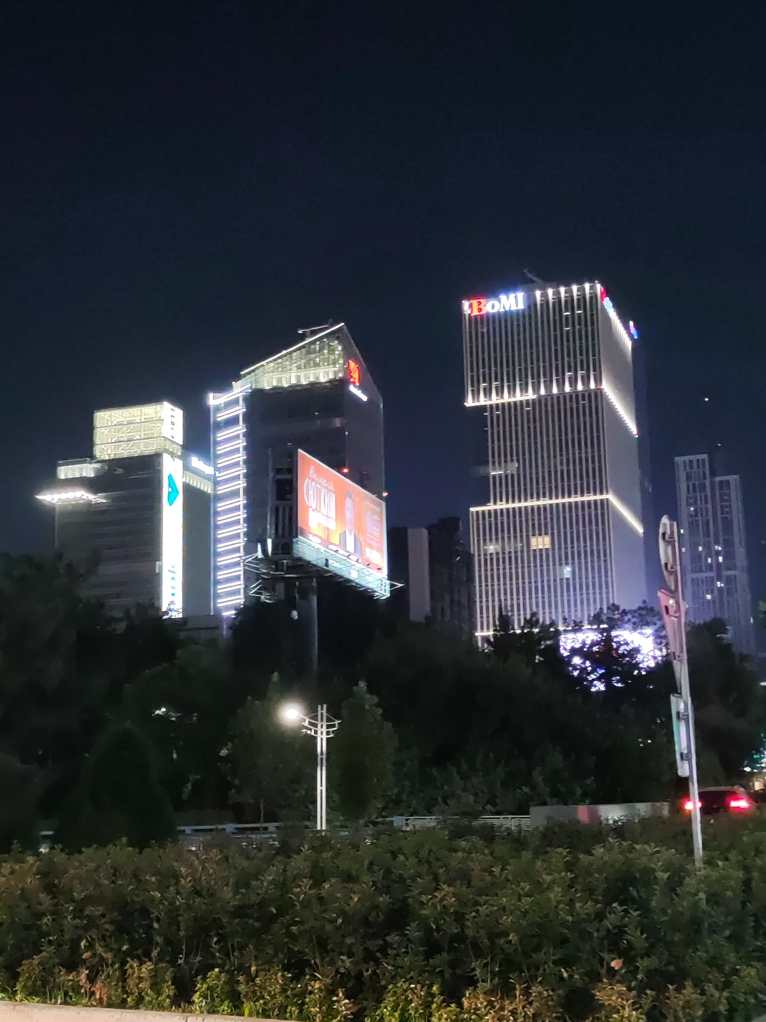 The image shows a nighttime view of a modern cityscape with several illuminated skyscrapers. One prominent building displays the "BOMi" logo in red and blue lights at its apex. Another building, to the left, has a large, illuminated billboard featuring a man's portrait and text in a foreign language, possibly Russian given the likely location. Trees and bushes form a dense foreground, with a street lamp visible on the right side of the image. A car with illuminated taillights is also visible in the distance. The architecture suggests a developed urban area. This scene is characteristic of the business district in Tashkent, Uzbekistan, specifically the area around Amir Timur Square, which features modern office buildings and prominent advertising.