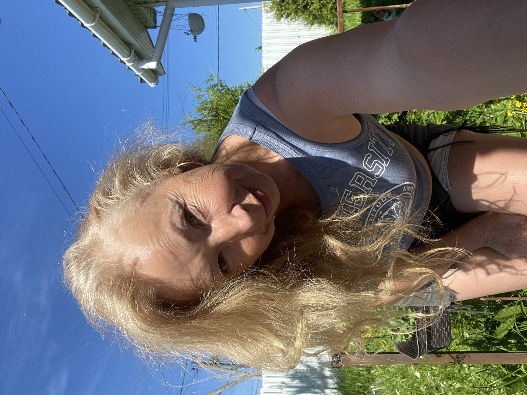 A blonde woman with fair skin and a light complexion, wearing a grey sleeveless crop top with "UNIVERSITY" written across it and denim shorts, is captured in a low-angle, selfie-style photograph outdoors under a clear blue sky. Her hair is styled in loose waves that fall across her face and shoulders. She is leaning forward, her face tilted towards the camera, with a slight smile. The background includes a white picket fence, green grass, bushes, and the corner of a white house with a gutter. The scene suggests a casual, sunny day in a residential area, likely in the city of Bol'shoe Zaborod'e, Russia.