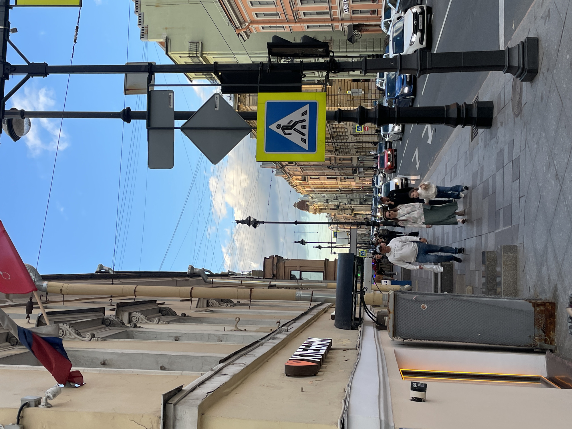 The image depicts a street scene in Saint Petersburg, Russia, likely on a sunny day given the bright blue sky with scattered clouds. The perspective is from a sidewalk looking down a street lined with buildings.
On the left, a building with a pale yellow facade features architectural details and a sign that appears to be in Cyrillic, with an orange logo. A Russian flag is partially visible, attached to the building. A street lamp with a globe-shaped fixture stands tall on the right side of the frame, part of a decorative black metal lamppost that also supports various traffic signs.
In the foreground, a paved sidewalk is occupied by several people. A man in a white sweatshirt and jeans walks with a plastic bag in his hand, accompanied by a woman in a green skirt and a patterned top, and a young girl. They are walking away from the camera, towards the other pedestrians and traffic further down the street.
The street itself is busy with vehicular traffic, including cars of various colors. Traffic signs are visible, including a pedestrian crossing sign. The buildings lining the street are of varying architectural styles and colors, suggesting a historic urban setting. The overall impression is of a lively, urban environment in Saint Petersburg.