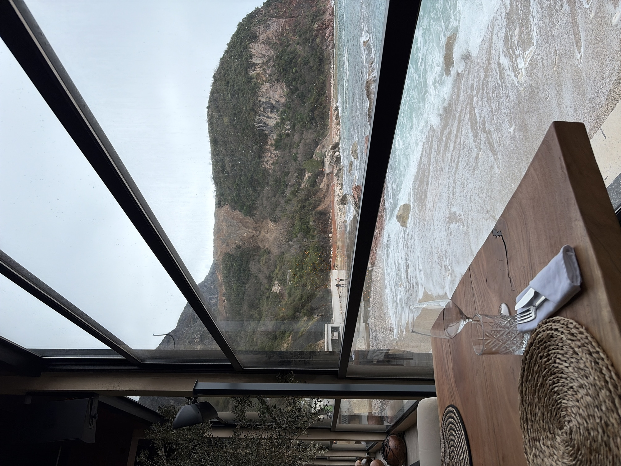 The image depicts a view from a commercial establishment, likely a restaurant or cafe, looking out through large, multi-paned windows onto a dynamic coastal scene within the city of Unknown, Unknown.
**Interior (Foreground):** A polished wooden table, featuring distinct grain, occupies the lower right quadrant. On the table, an empty stemmed wine glass and an empty patterned drinking glass stand beside a set of silverware (fork and knife) neatly folded within a light grey napkin, all positioned on a circular woven placemat. A second, partially visible woven placemat is present further back on the left. To the far left, parts of the interior infrastructure are visible: a dark structural column, a speaker mounted high, and the foliage of a live plant, possibly an olive tree. Beige upholstered seating is partially visible behind the table on the left. The window frames are dark, likely black or dark brown.
**Exterior (Midground/Background):** Beyond the windows, a pebbled and sandy beach stretches along the coast, where white-foamed waves crash onto the shore. The sea, appearing choppy, displays shades of turquoise and grey. In the mid-distance, two individuals are standing on a light-colored concrete platform or pier that extends slightly into the water from the coastline. They are observed from behind, facing the sea. The coastline rises steeply into a large, vegetated hillside featuring sections of exposed reddish-brown rock and areas of disturbed earth, possibly from erosion or construction. A small structure with a light-colored roof is visible near the base of the hillside. A narrow dirt track runs along portions of the hillside above the beach. The sky above is overcast and uniformly light grey.