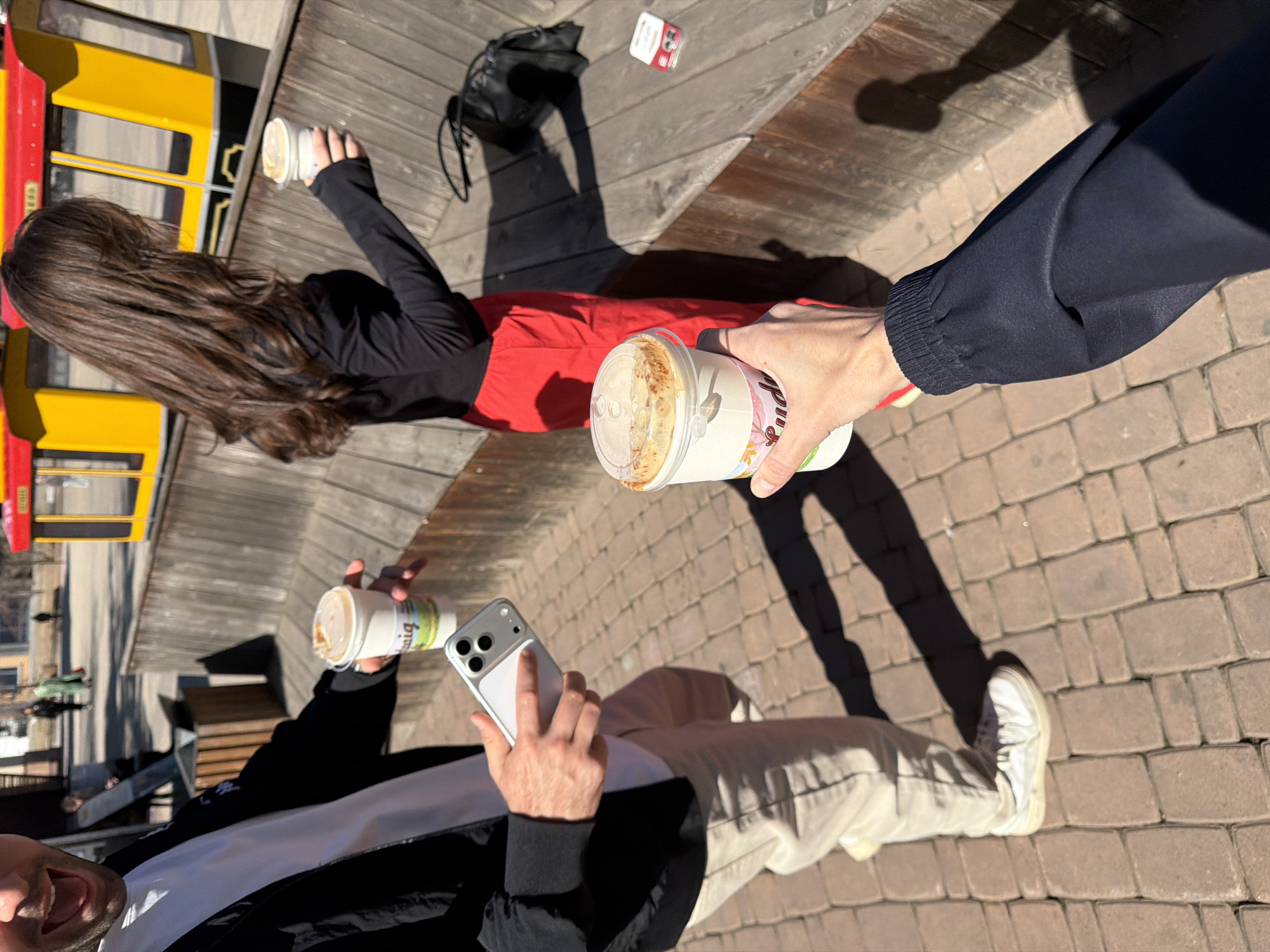 This image depicts an outdoor social gathering of at least three individuals on a sunny day in Krasnoyarsk, Russia, viewed from a slightly elevated perspective looking down.
In the foreground left, a person (likely male) is partially visible, showing the top of their head with an open mouth, suggesting they are talking or smiling. They wear a black jacket, a white undershirt, and light-colored pants. This person holds a smartphone (appearing to be an iPhone with a silver case and triple cameras) in their left hand and a white coffee cup, partially revealing the text "könig," in their right.
To the right in the foreground, another person's arm and hand are extended towards the center, holding a coffee cup with a colorful wrapper that shows "Puddi..." (likely "Pudding") text. They wear a dark sleeve.
Further back and to the left, a third person, a female, is seen from behind with long, dark, wavy hair. She wears a black long-sleeved top and red pants, sitting or leaning on a wooden deck. She holds a coffee cup in her left hand, while her right arm is outstretched. Nearby on the wooden deck are a black bag and a small white box with "Tic Tac" visible on its red label.
The ground consists of wooden decking in the background and light-colored cobblestone pavers in the foreground. The bright sunlight casts distinct, long shadows, indicating either morning or late afternoon. In the background, a prominent yellow and red structure, possibly a kiosk or part of a building, is visible, alongside other distant urban elements and trees. The scene captures a relaxed social moment, with individuals enjoying beverages outdoors.