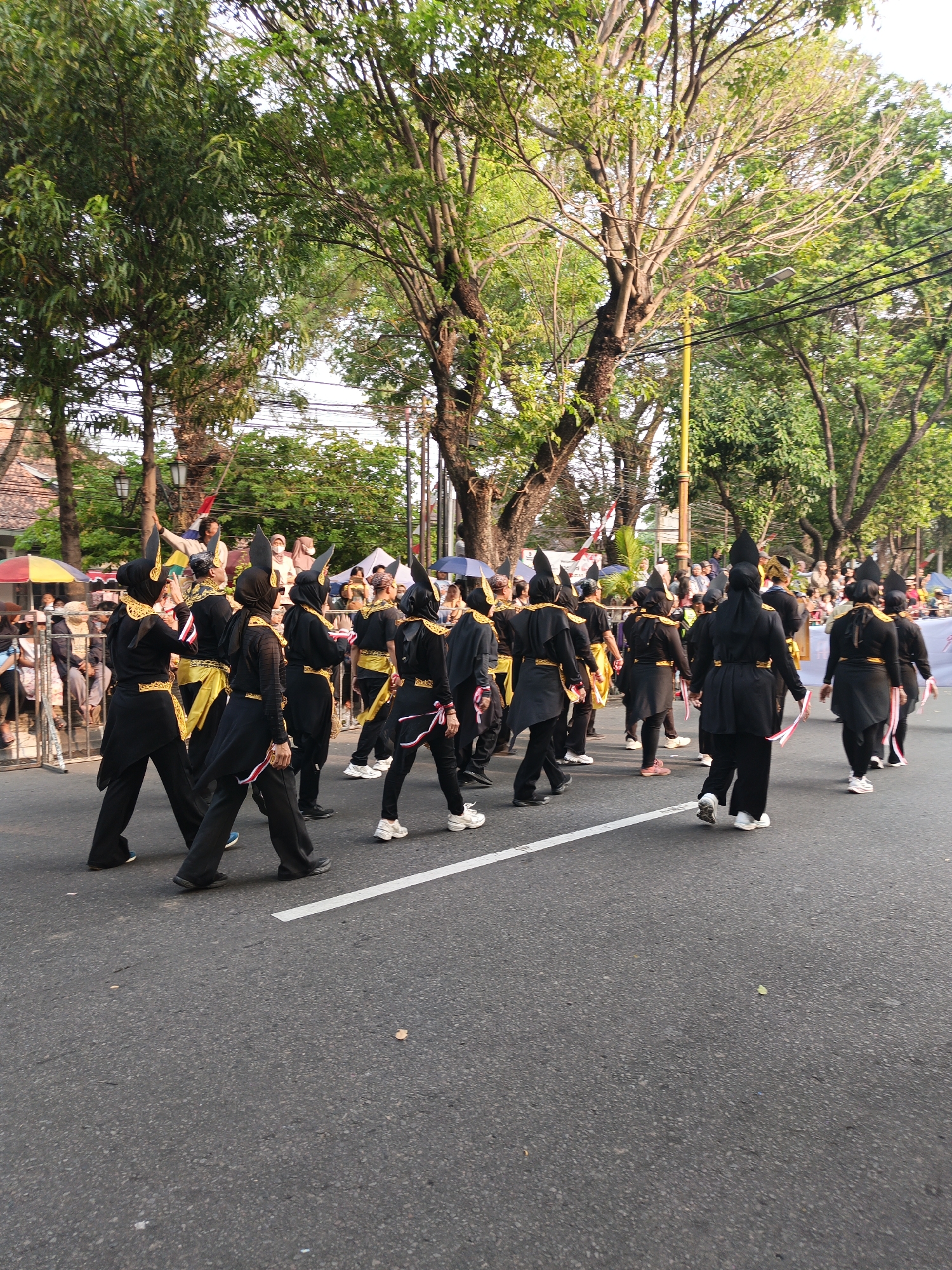In the city of Gayamprit, Indonesia, a group of people in matching black and gold traditional costumes are marching down a paved street. The costumes feature prominent, pointed headwear and gold sashes. The individuals are walking in unison, facing away from the camera, and some are carrying small red and white flags. They are on a road with a white painted line on the right side. The background consists of trees with lush green foliage and a clear sky, suggesting it is daytime. There are spectators visible in the background on the left side, some seated and others standing, behind a metal barrier. Additionally, there are several tents with colorful awnings and streetlights present in the scene.