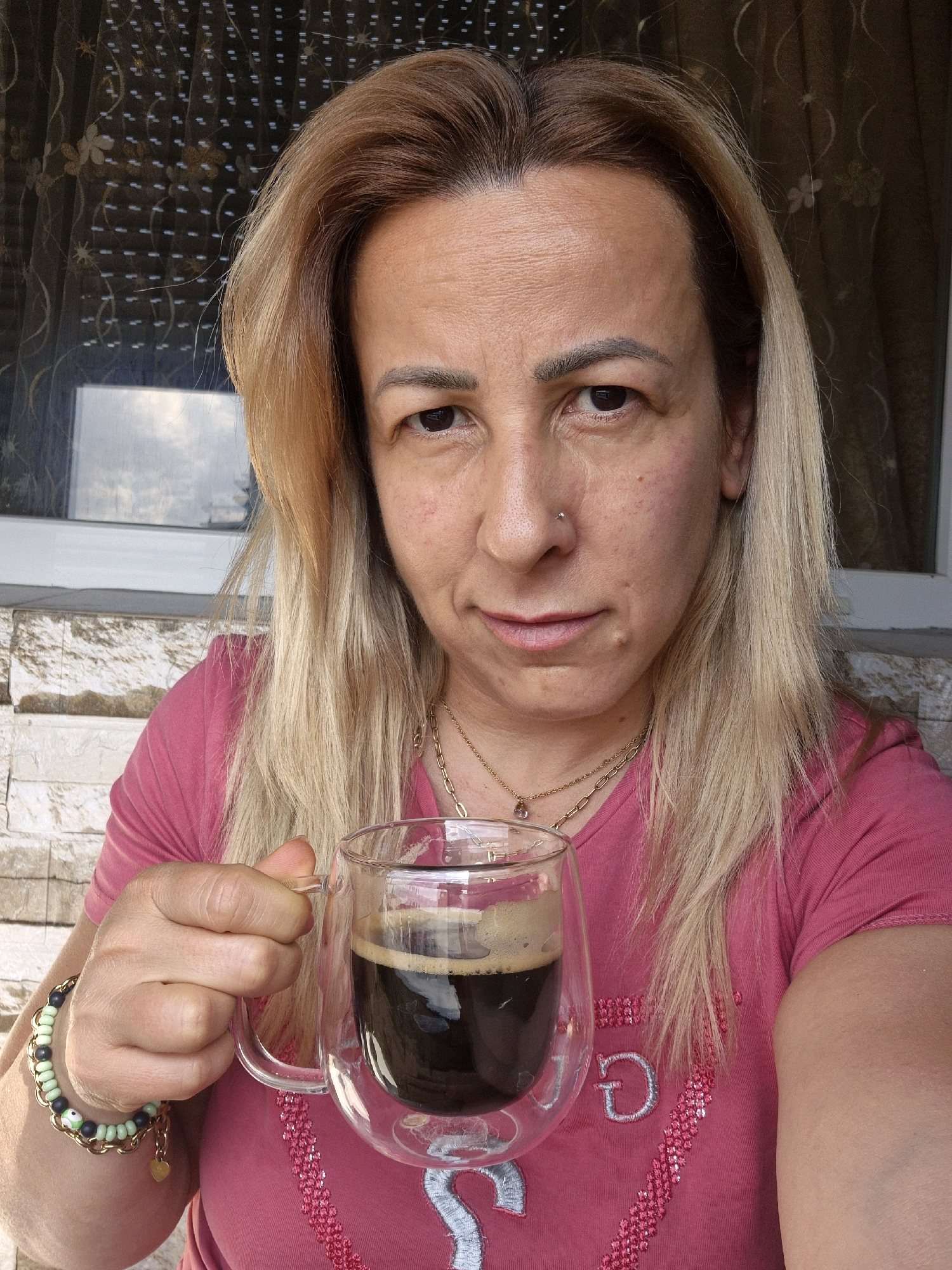 A woman with blonde hair and fair skin is holding a glass mug of coffee. She is wearing a pink t-shirt with silver embellishments. She has a nose ring and several necklaces. The background appears to be an outdoor setting with a brick wall and a window. The image is a selfie.