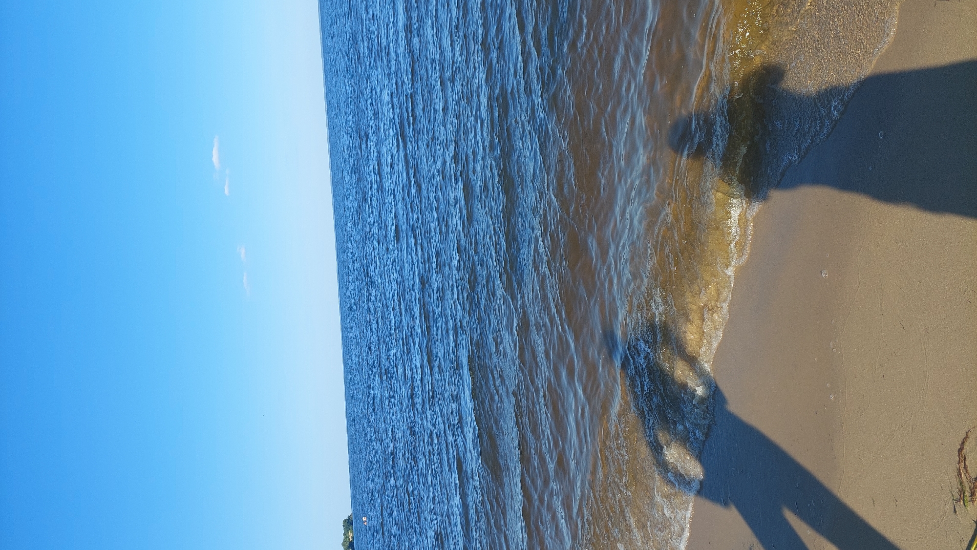 The image depicts a wide, calm body of water, likely a lake or a large river, under a clear blue sky with a few wispy clouds. The water's surface is gently rippled and has a brownish hue where it meets the shore, suggesting shallow, sandy bottom. The foreground is a sandy beach with two distinct shadows cast by individuals, one larger and to the left, the other smaller and to the right. The shadows suggest the presence of people on the beach, possibly taking photos or enjoying the view. In the distance, on the left side of the frame, a strip of green land with some trees is visible, hinting at a shoreline or an island. The overall scene conveys a sense of tranquility and a typical summer day at a waterfront location in Kozarovychi, Ukraine.