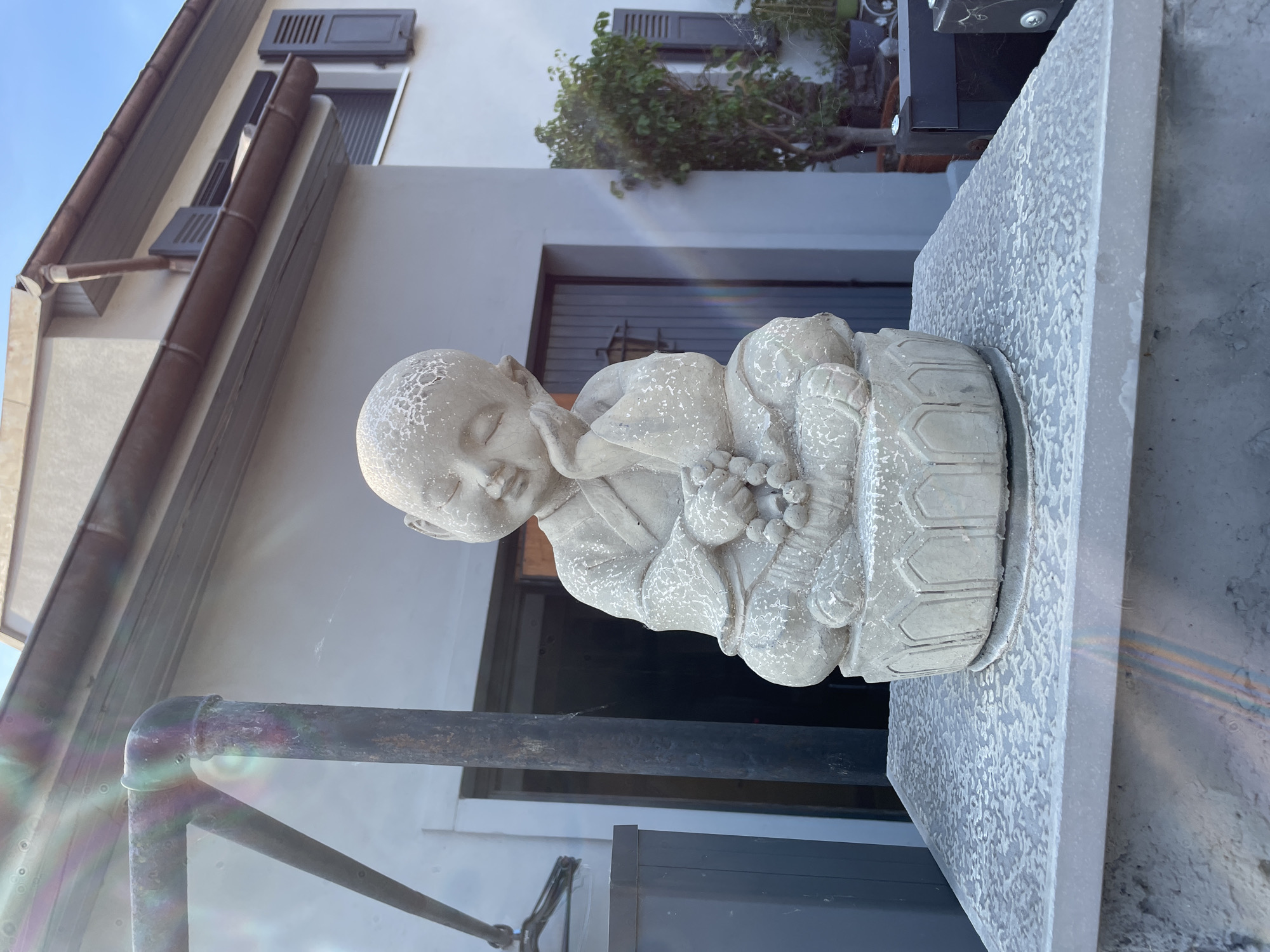 A concrete statue of a young meditating monk is positioned outdoors on a stone pedestal. The monk statue is seated cross-legged, with its right hand supporting its head as if in deep thought or sleep, and its left hand resting on a string of beads. The statue has a serene expression and is wearing traditional monk attire. The pedestal is light gray and textured with a pattern of irregular white spots, resembling a distressed or weathered finish.
Behind the statue, a building with light gray walls and brown shutters on the windows is visible. A dark-colored door is also present on the building. To the right of the statue and building, a green tree in a pot adds a touch of nature to the scene. The sky above is a clear blue. The overall environment suggests an outdoor setting, possibly a garden or courtyard, with a residential or commercial building in the background. The image exhibits lens flare with rainbow-like refractions, particularly noticeable in the upper left and lower portions. The location is identified as Gerstheim, France.