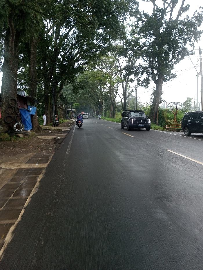A wide-angle, eye-level outdoor shot depicts a two-lane asphalt road, visibly wet, in YonPhoogla, Bhutan. The road is dark grey with a yellow dashed line marking the center and a white solid line indicating the left edge. Lush green foliage from numerous large, mature trees forms a dense canopy overhead, particularly in the distance.
On the left side of the road, a concrete sidewalk with a textured pattern leads to a dirt shoulder. A makeshift stall or shelter, constructed with a blue tarp, is affixed to the base of a large tree trunk. This stall is adorned with several black rubber rings or tire rims and a white circular object. A person in a long white garment stands partially obscured by a tree near this stall. Further down the left shoulder, a red motorcycle is present. In the left lane, a blue scooter ridden by a single individual wearing a dark helmet and dark clothing travels away from the viewer. A white van is also visible further in the distance, moving in the same direction.
On the right side of the road, a dark-colored SUV, resembling a Mitsubishi Xpander Cross, with a roof rack and square headlights, travels towards the viewer in the right lane. Its front license plate is partially visible, beginning with "DK." Behind the SUV, a dark-colored minivan or SUV is parked partially off the road on a grassy shoulder. The right side is also lined with large trees, and a light-colored, possibly wooden, framework structure with a yellow base is visible amidst the greenery in the mid-ground. Utility poles with overhead wires are present on both sides of the road. The sky is overcast and light grey, visible through gaps in the tree canopy.