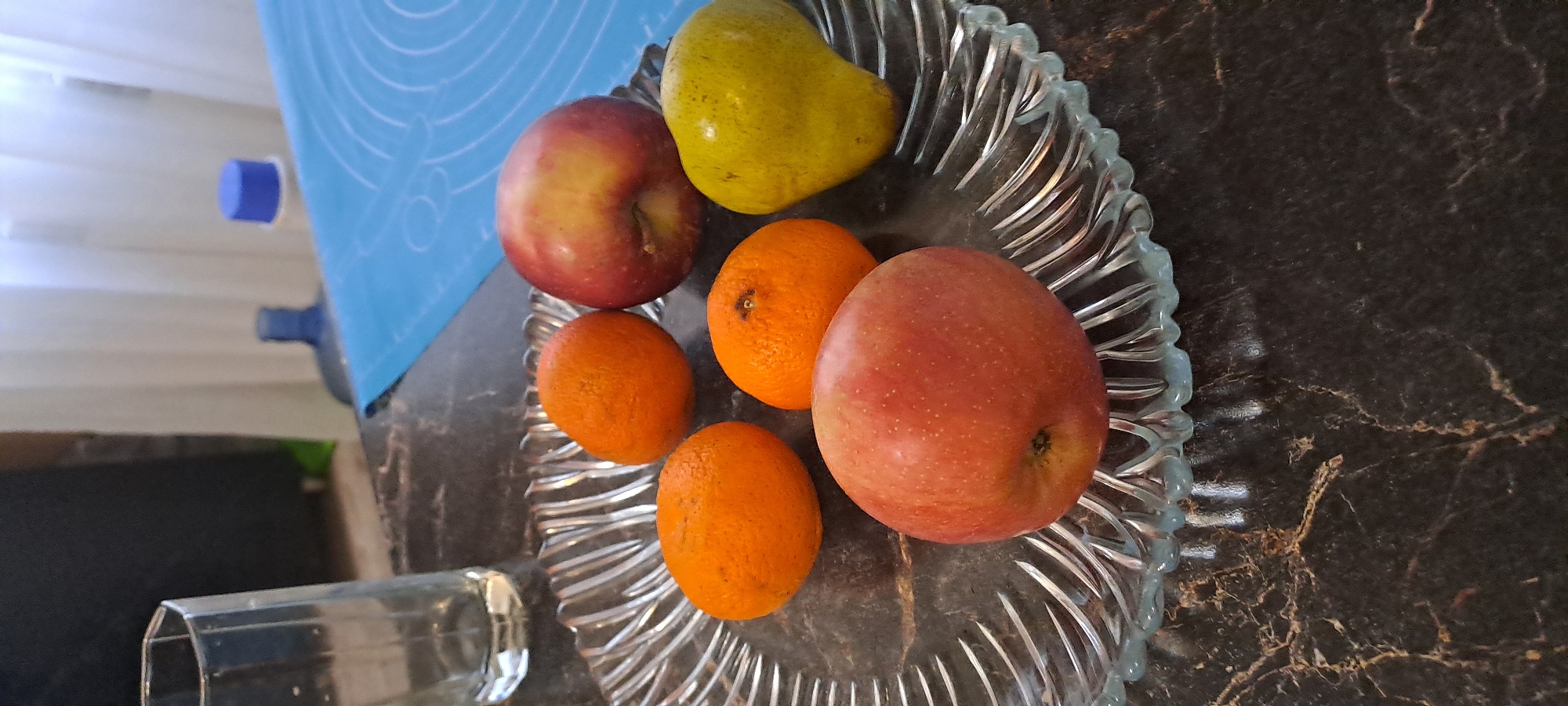 The image shows a glass plate containing two apples, three oranges, and a pear. The plate sits on a dark, patterned surface. To the left of the plate, there is a glass and a blue object in the background. The image does not contain information to identify the location in Yekaterinburg, Russia or any people, actions, or interactions.