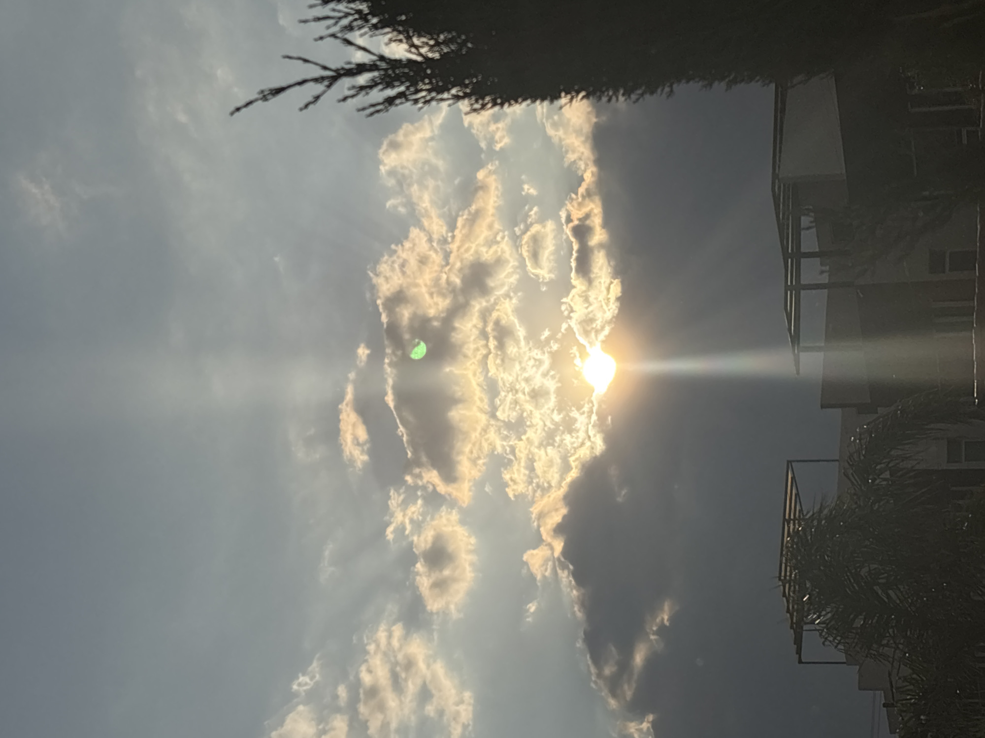 The image shows a view of the sky with bright sunlight shining through clouds. The sun is mostly obscured by the clouds, but beams of light radiate outwards. Part of a building is visible on the right edge of the frame, with some palm fronds partially obscuring it. Some tree branches are at the top of the frame. The photo appears to have been taken in San Juan Cuautlancingo, Mexico, as specified.