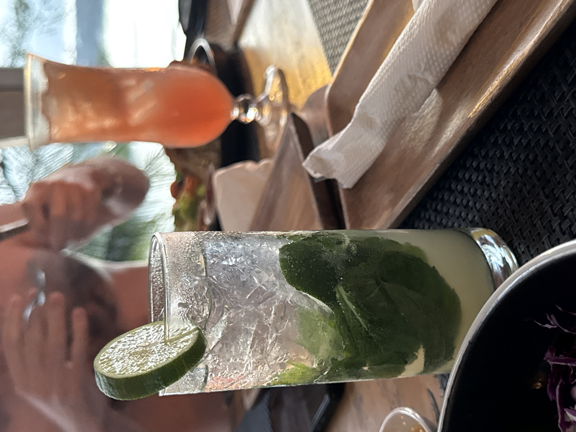 An outdoor table at a restaurant in Nusa Penida, Indonesia, is set for a meal. In the foreground, a tall, clear glass filled with ice, mint, and a lime slice is a mojito. To the right of the mojito, a wooden tray holds a white paper napkin. Further in the background, another tall glass with a red drink sits on the table, and a person's hands are visible above it, holding cutlery, suggesting they are eating. The environment appears to be a tropical setting with hints of greenery in the background, typical of Nusa Penida.
